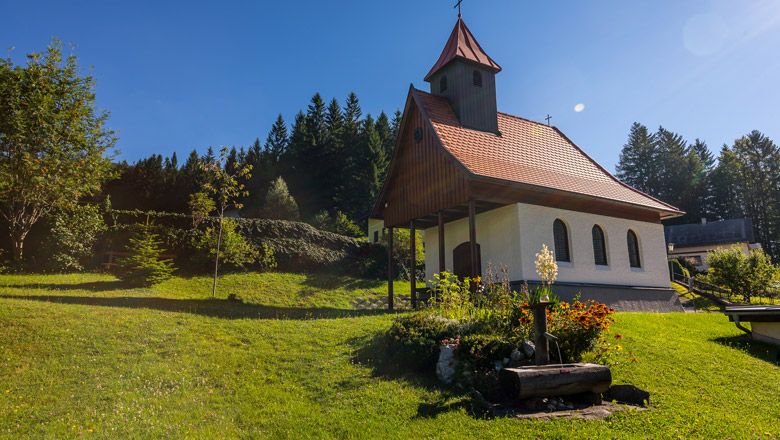 Malá kaplnka s červenou strechou na zelenej lúke, obklopená stromami a kvetmi.