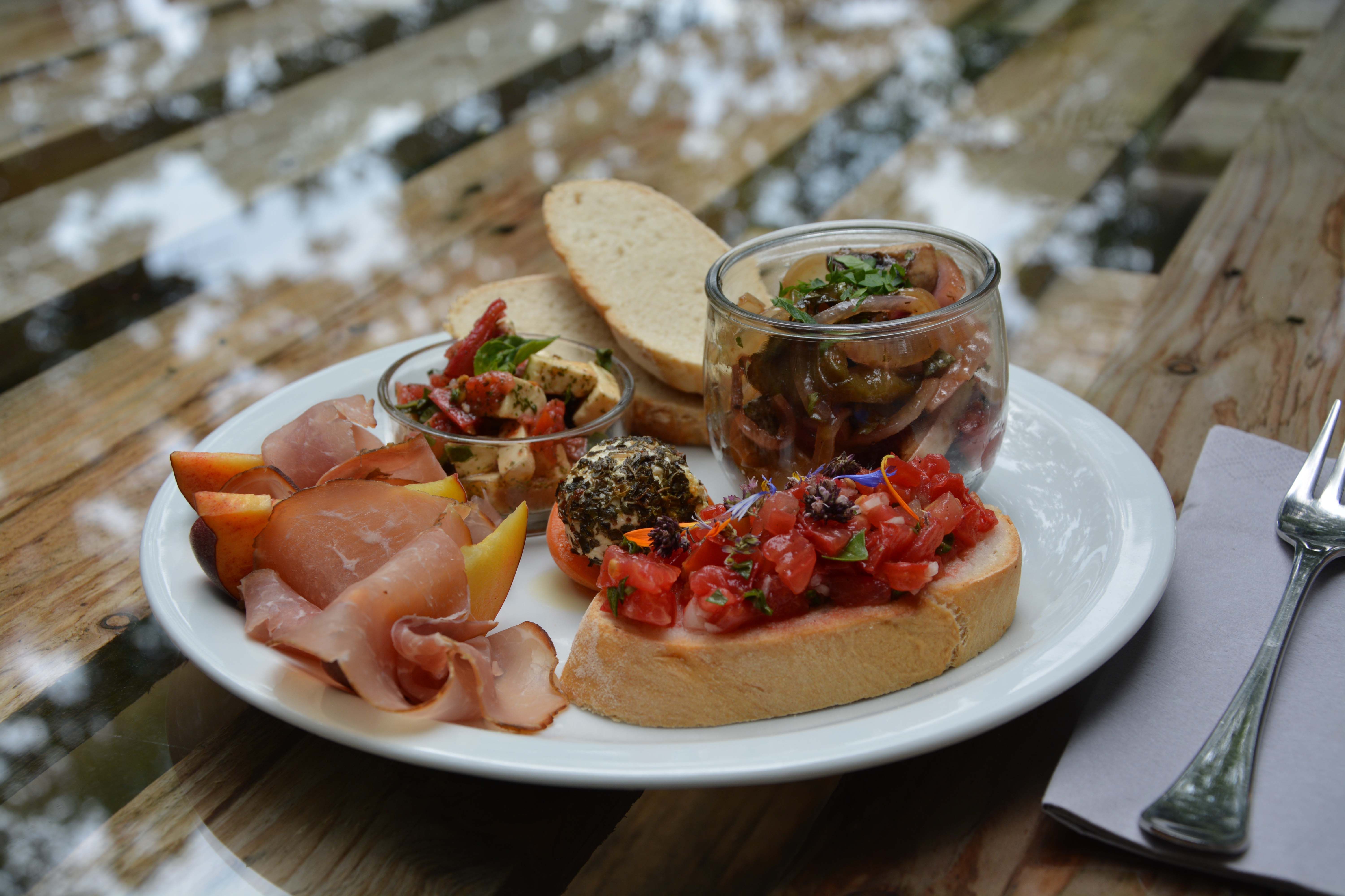 Na drevenom stole tanier s chlebom, šunkou, antipasti, bruschetou a ovocím.