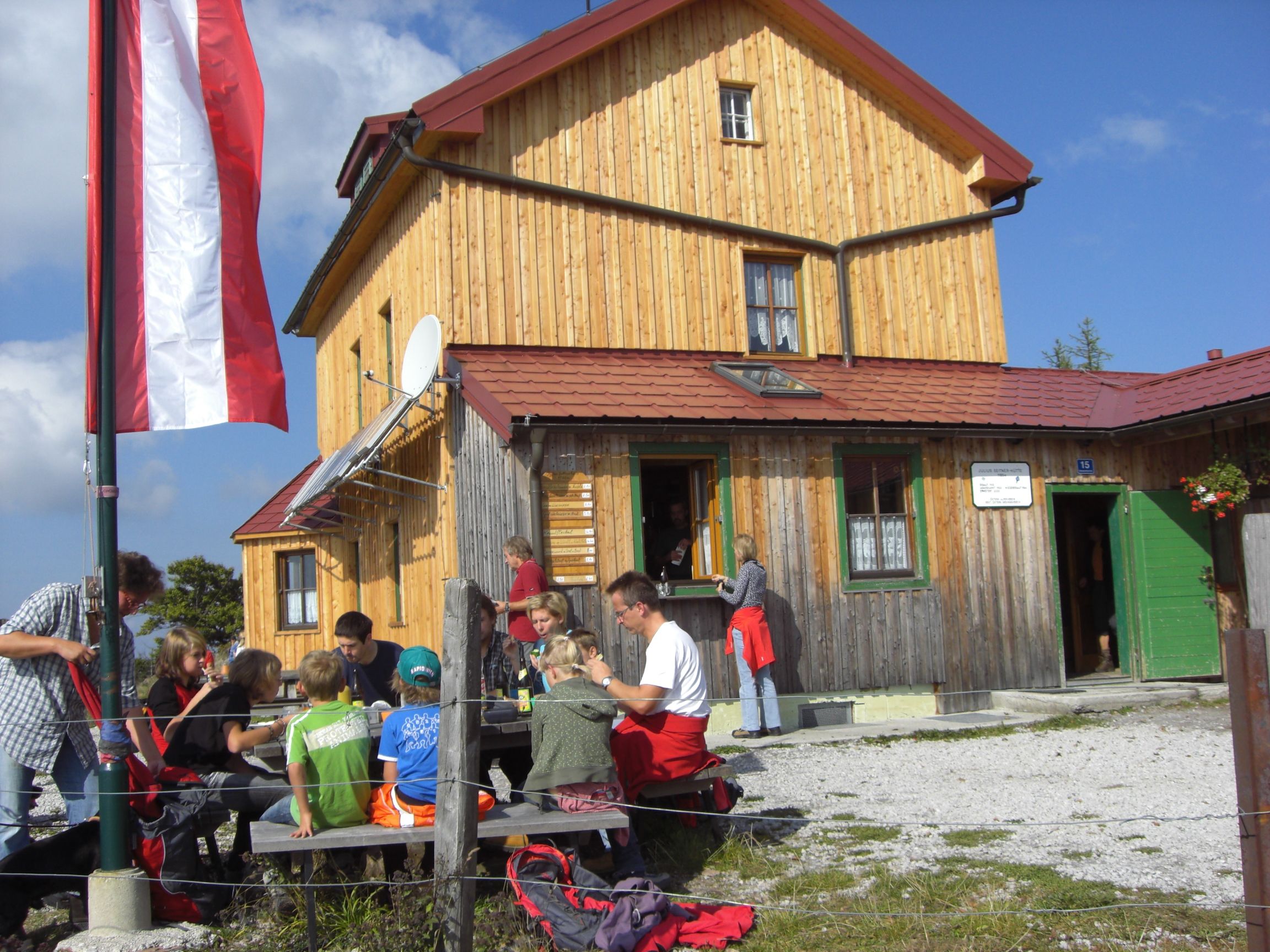 Skupina ľudí sedí pred horskou chatou s drevenou fasádou a rakúskou vlajkou.