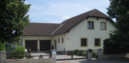 Moderný rodinný dom s hnedou strechou a svetlým náterom, obklopený stromami a plotom.