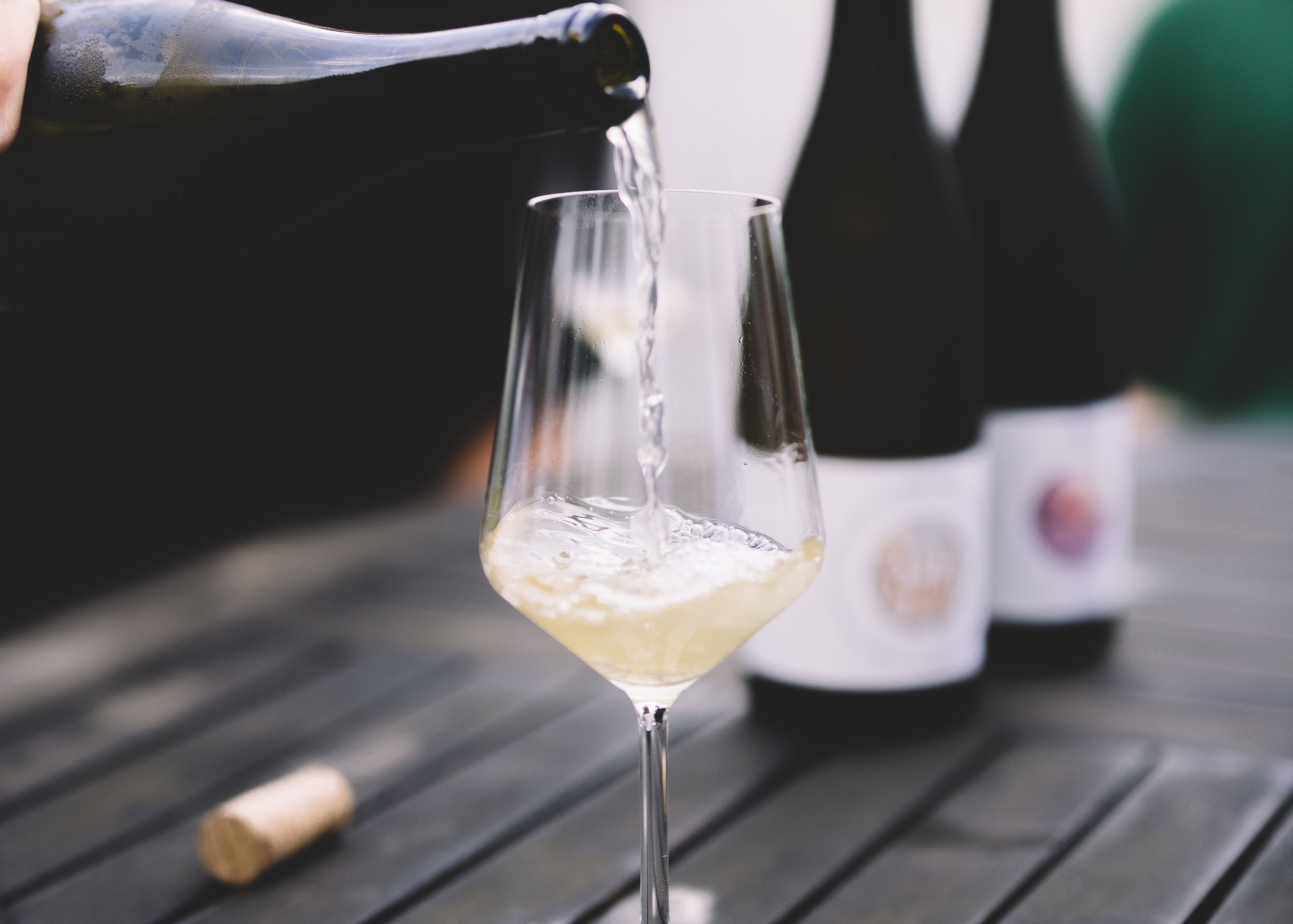 Na stole je naliaty pohár bieleho vína, v pozadí sú dve fľaše vína.