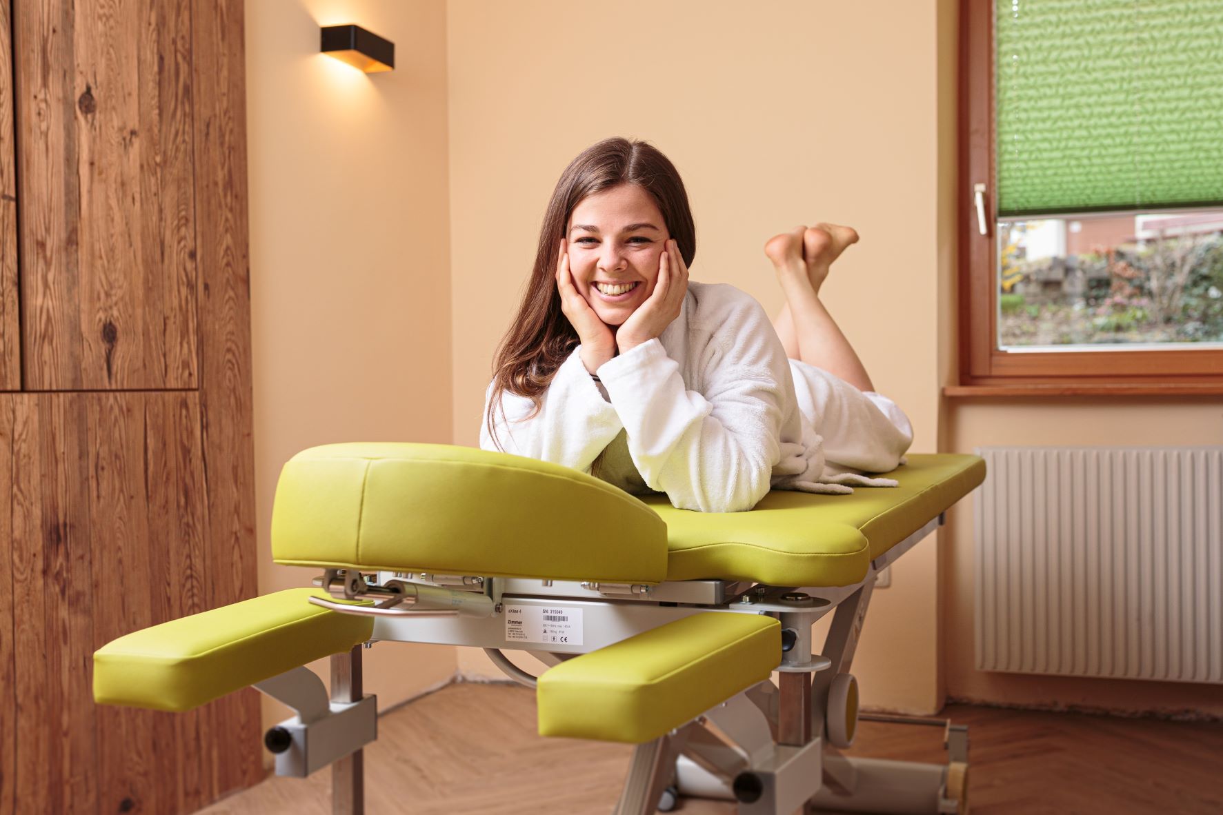 Žena v župane leží s úsmevom na zelenom masážnom stole vo wellness miestnosti.
