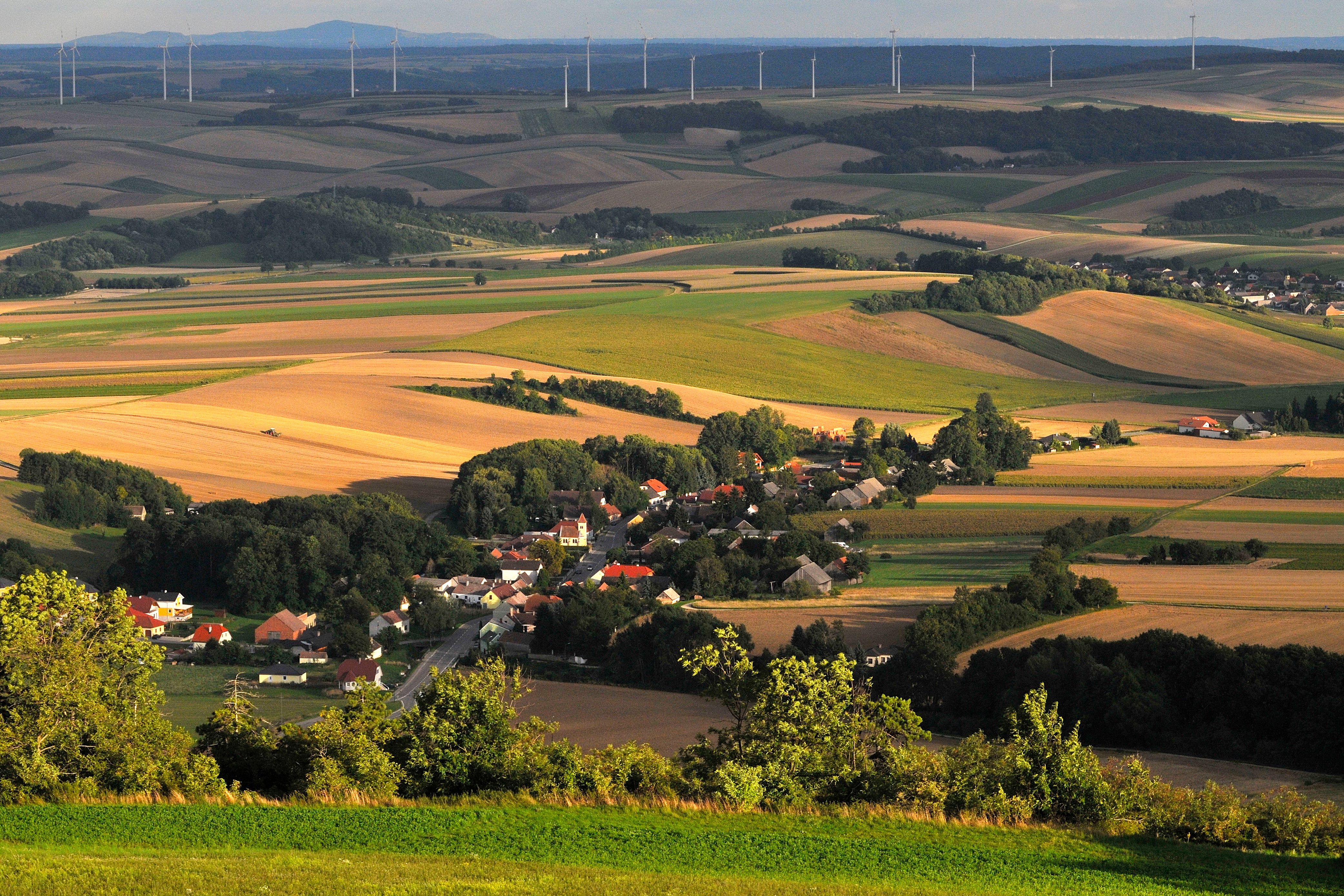 Krajina s poliami, dedinou a veternými turbínami v pozadí.