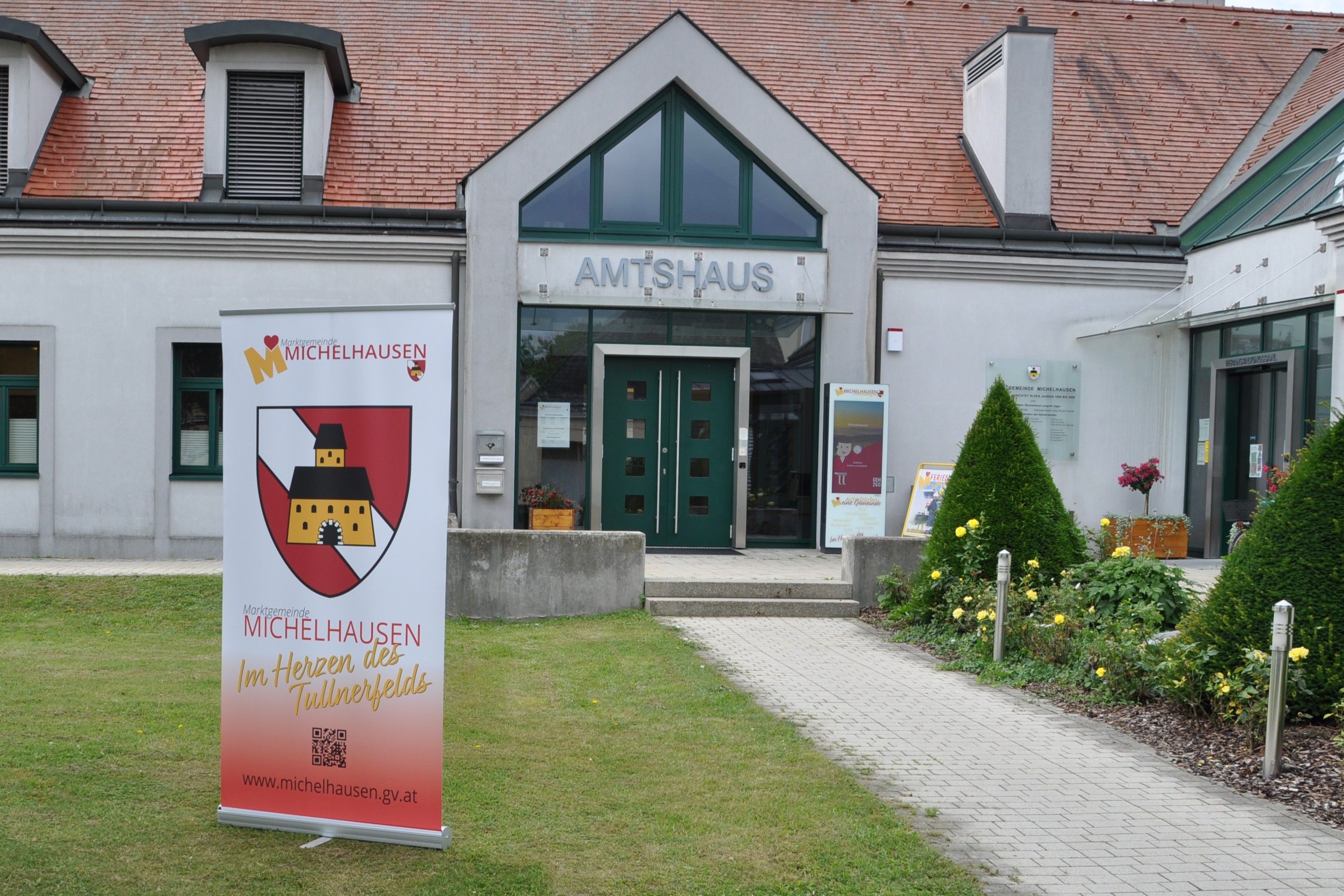 Vchod do domu Amtshaus Michelhausen s reklamným pútačom a udržiavanou záhradou.