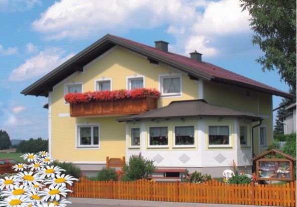 Žltý dom s červenou strechou a kvetinovou truhlicou na balkóne, obklopený dreveným plotom a sedmokráskami v popredí.