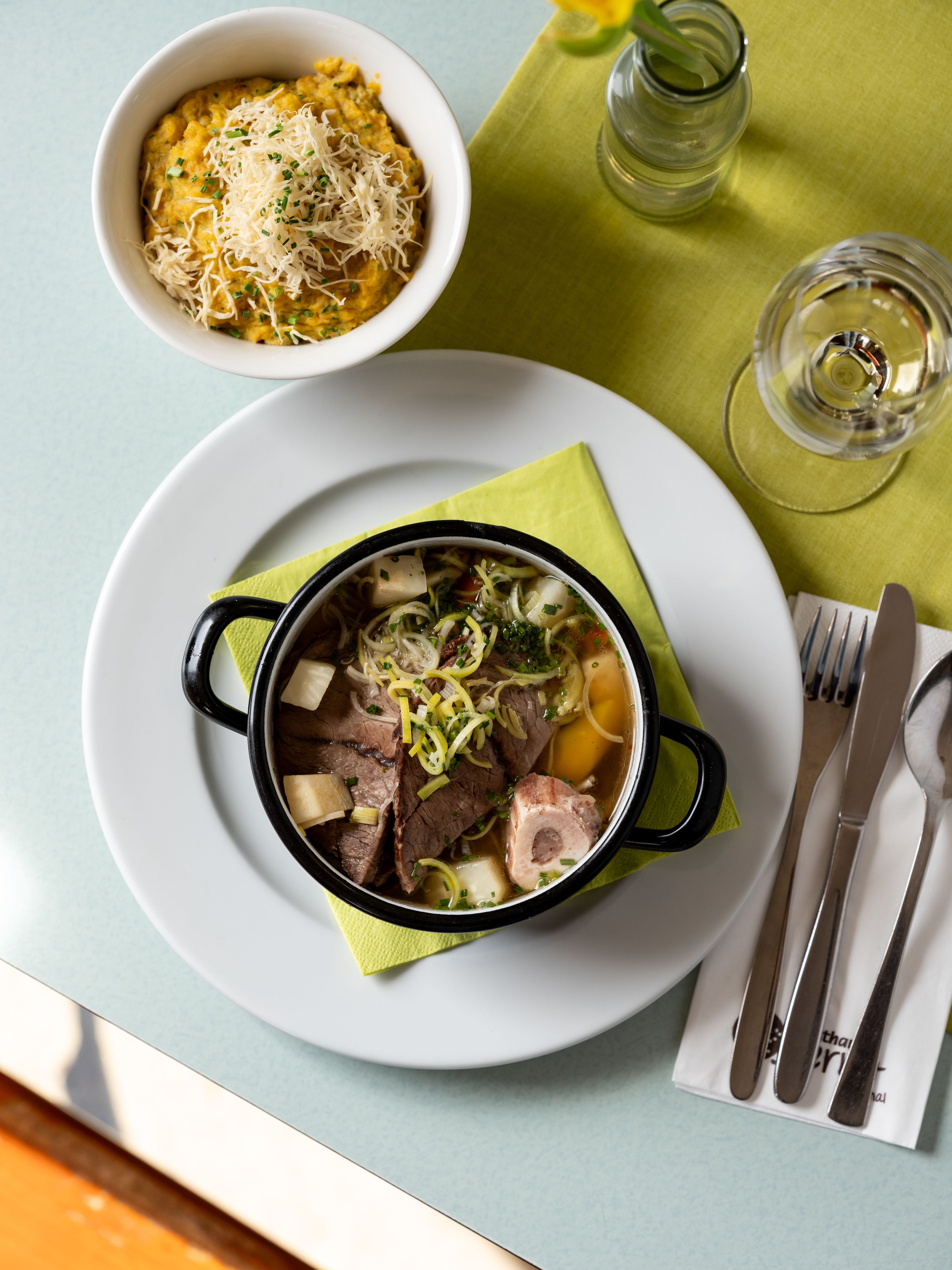 Stôl s tanierom polievky, miskou kaše so syrom, pohárom vody a príborom na zelenom obrúsku.