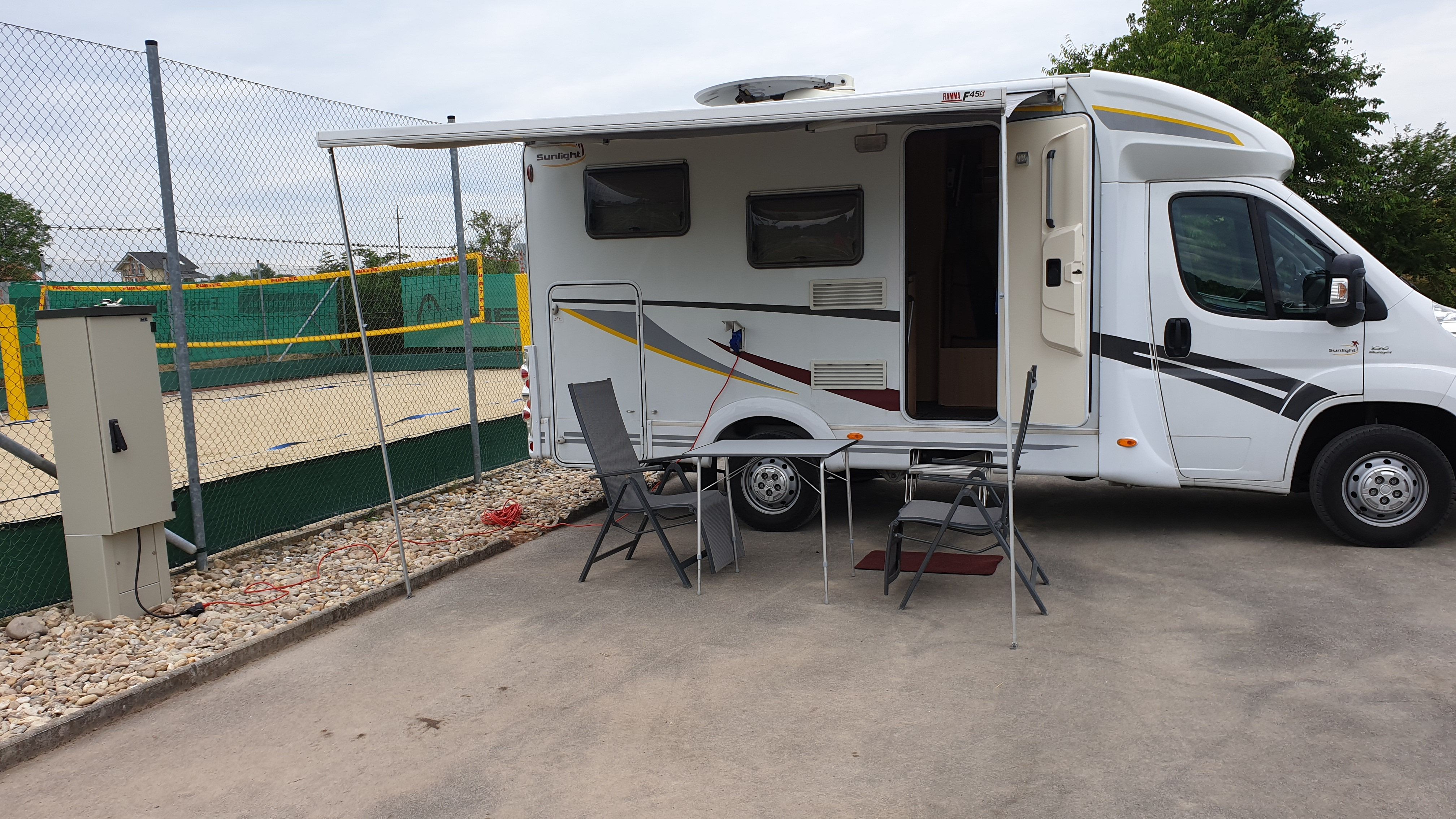 Karavan s predĺženou markízou a kempingovým nábytkom na pozemku vedľa volejbalového ihriska.