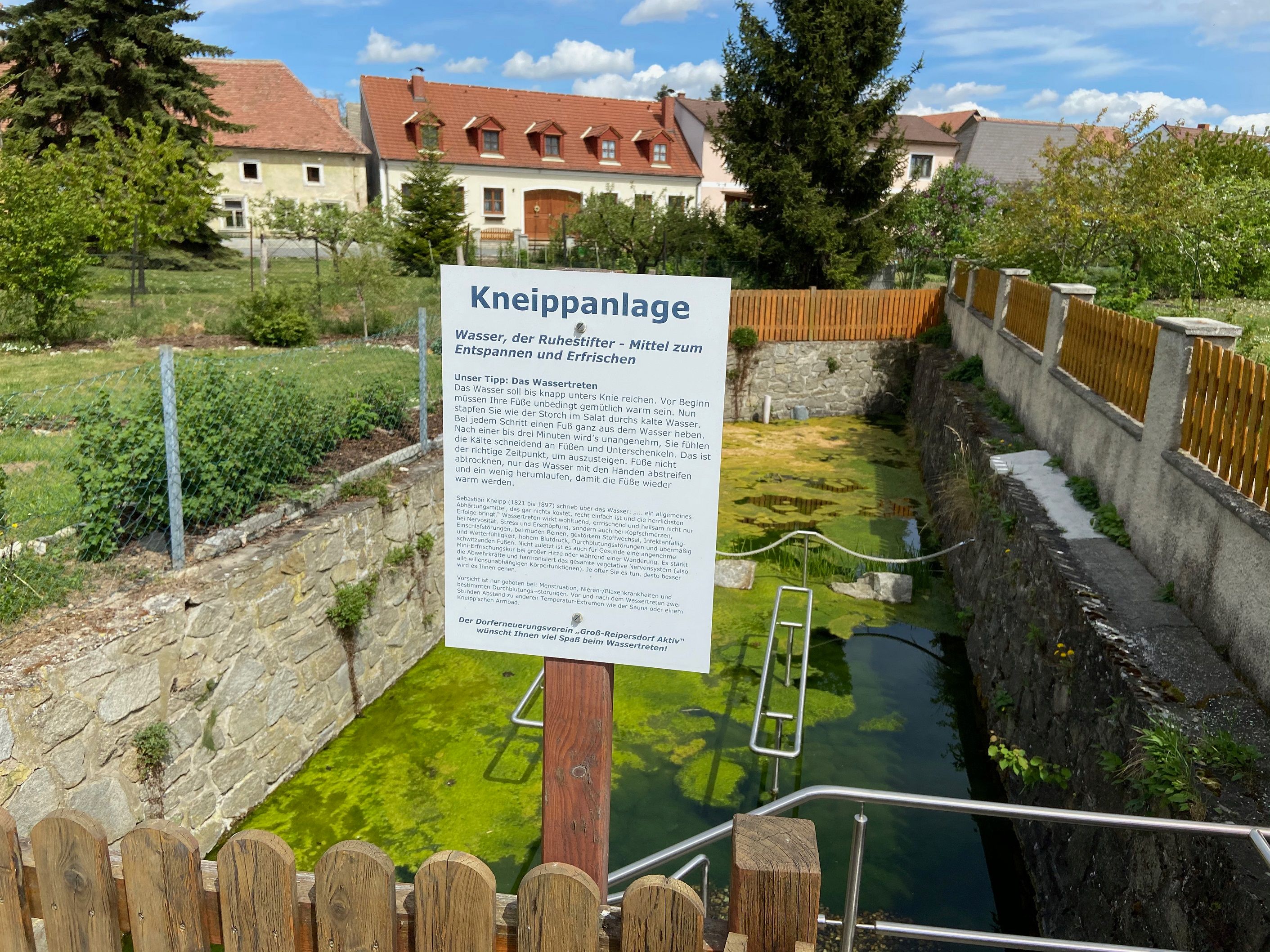Kneippovo zariadenie s informačnou tabuľou a nádržou na vodu, obklopené plotom a domami v pozadí.