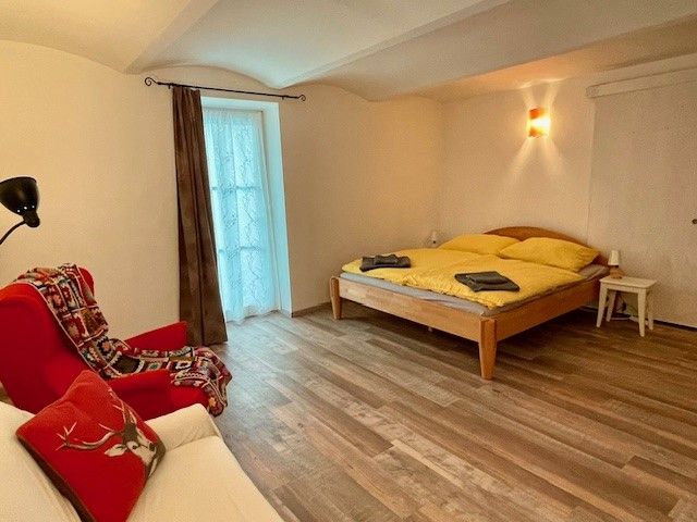 Útulná spálňa s drevenou podlahou, manželskou posteľou, červeným kreslom a oknom so závesom.