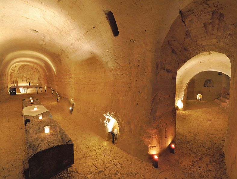 Podzemná pieskovcová pivnica so svetlom sviečok a rustikálnymi stolmi.