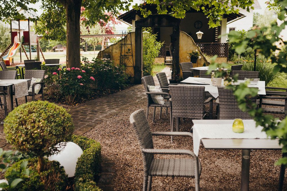 Útulný vonkajší priestor so stolmi a stoličkami pod stromami, obklopený rastlinami a detským ihriskom v pozadí.