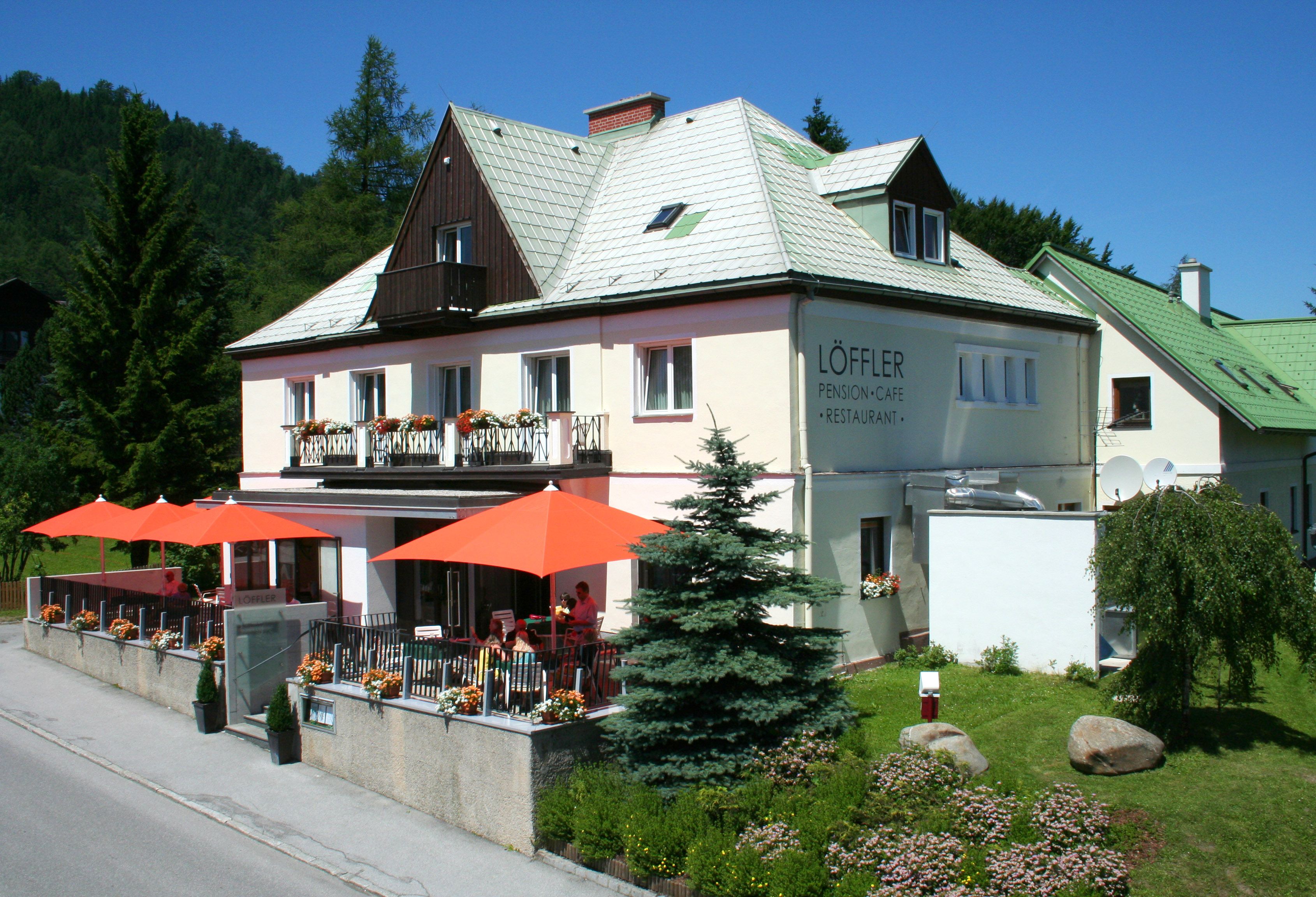 Dvojposchodová budova s označením "Löffler Pension-Café-Restaurant", obklopená stromami a kvetmi, s oranžovými slnečníkmi na terase.