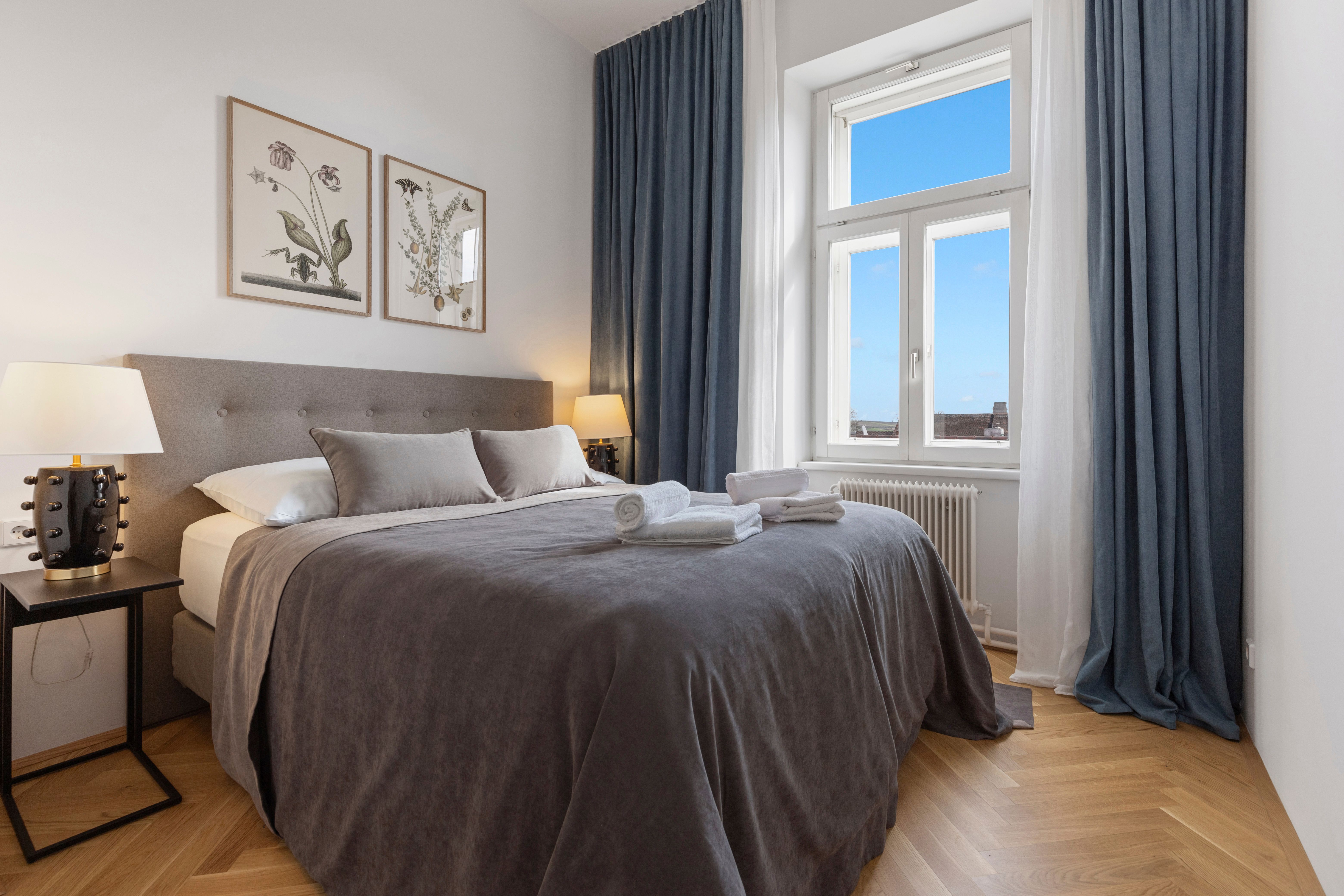 Elegantná spálňa s veľkou posteľou, sivou posteľnou bielizňou, dvoma nočnými stolíkmi s lampami a oknom s modrým závesom.