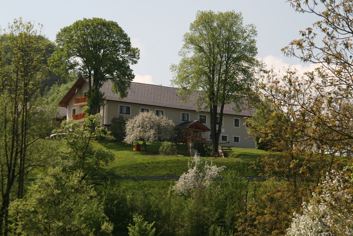Farma na zelenom kopci, obklopená stromami a kvitnúcimi kríkmi.