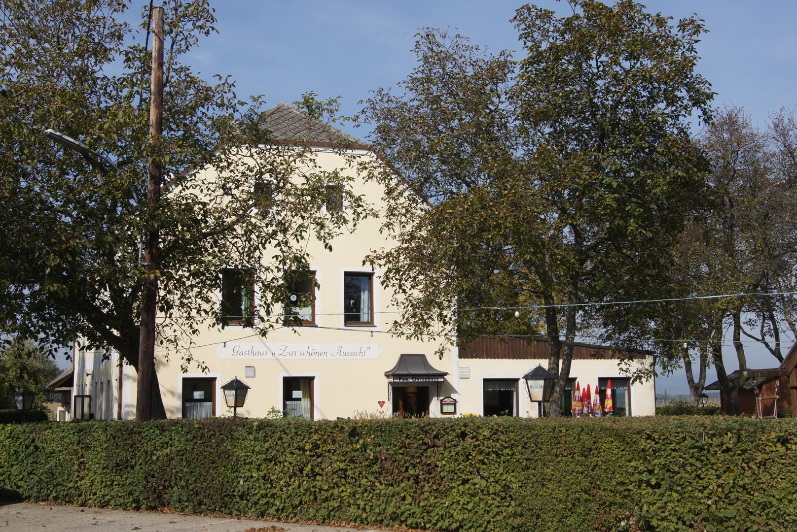 Žltý hostinec s nápisom "Gasthaus Zur schönen Aussicht", obklopený stromami a živým plotom.