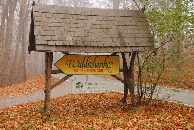 Drevená tabuľa v lese ukazuje na Waldschenke Holzkohlengrill.