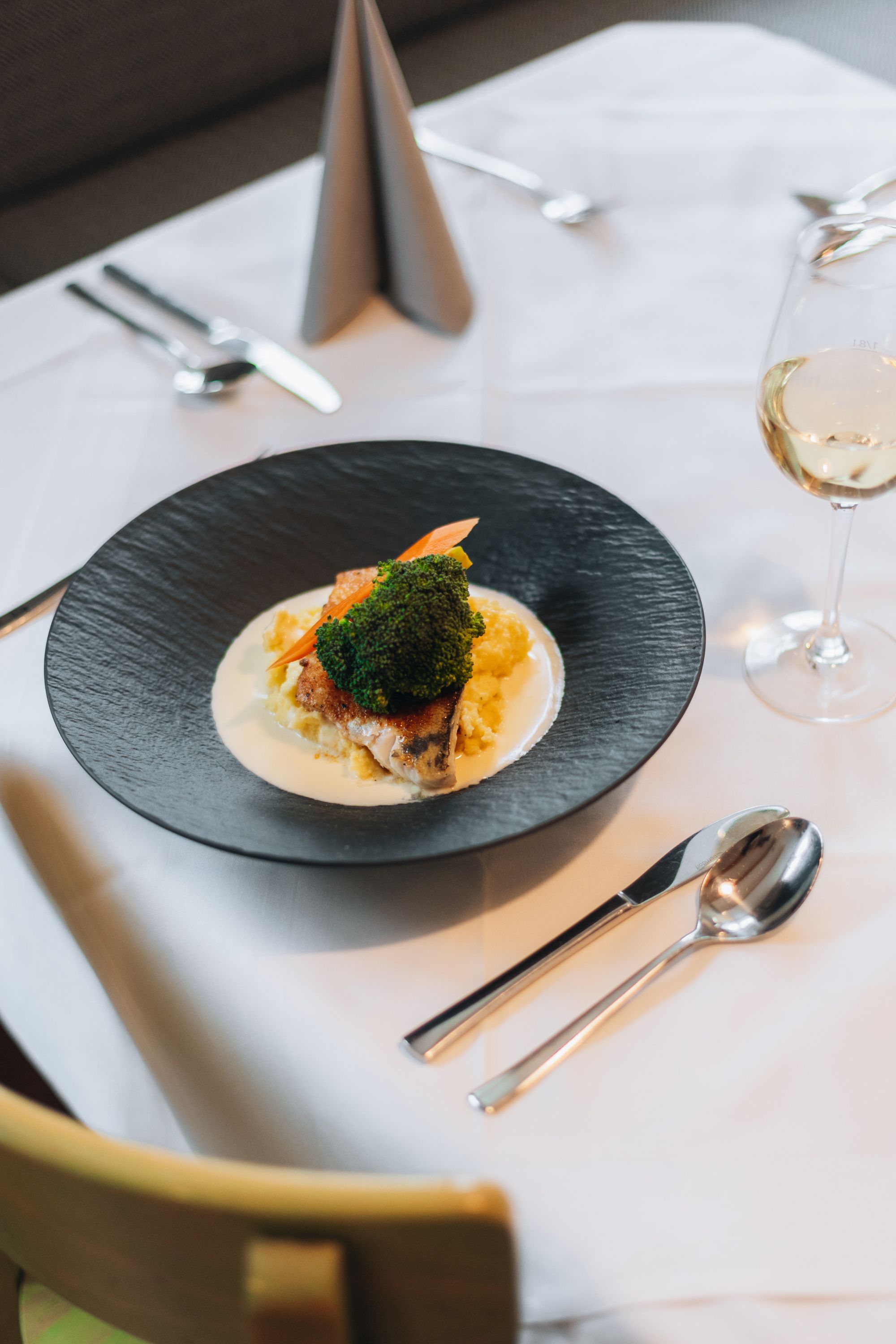 Elegantne prezentované jedlo na čiernom tanieri so zeleninou a omáčkou, doplnené pohárom bieleho vína na prestretom stole.