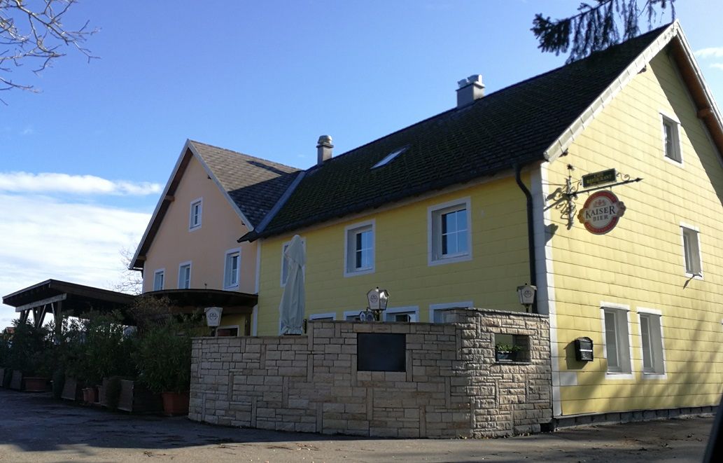 Žltý vidiecky hostinec s nápisom Kaiser Bier a slnečníkom v popredí.