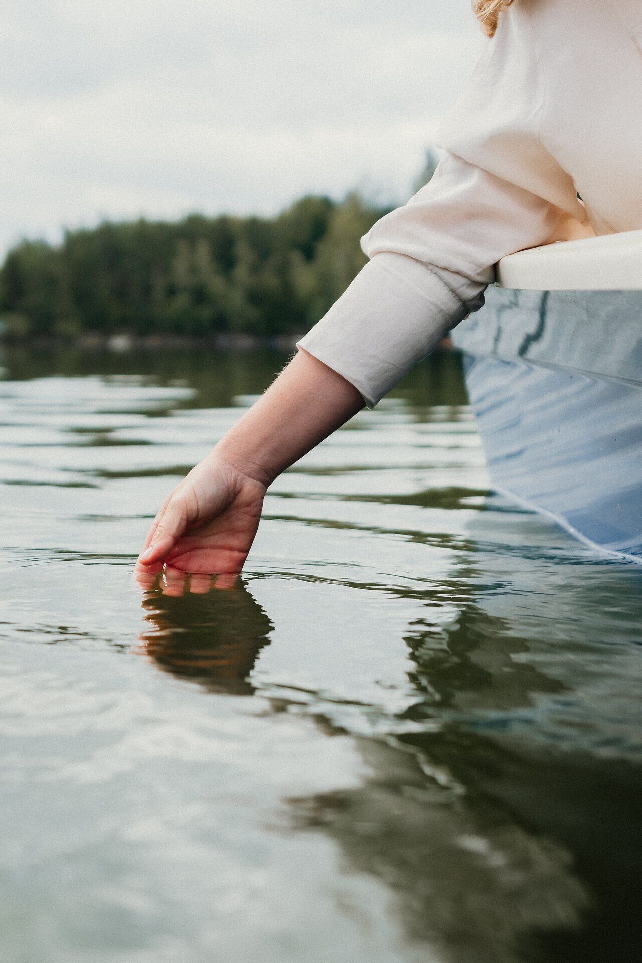 Hand einer Person taucht vom Rand eines Ruderboots ins ruhige Wasser des Stausees Ottenstein; im Hintergrund spiegeln sich Wald und Himmel auf der Wasseroberfläche.