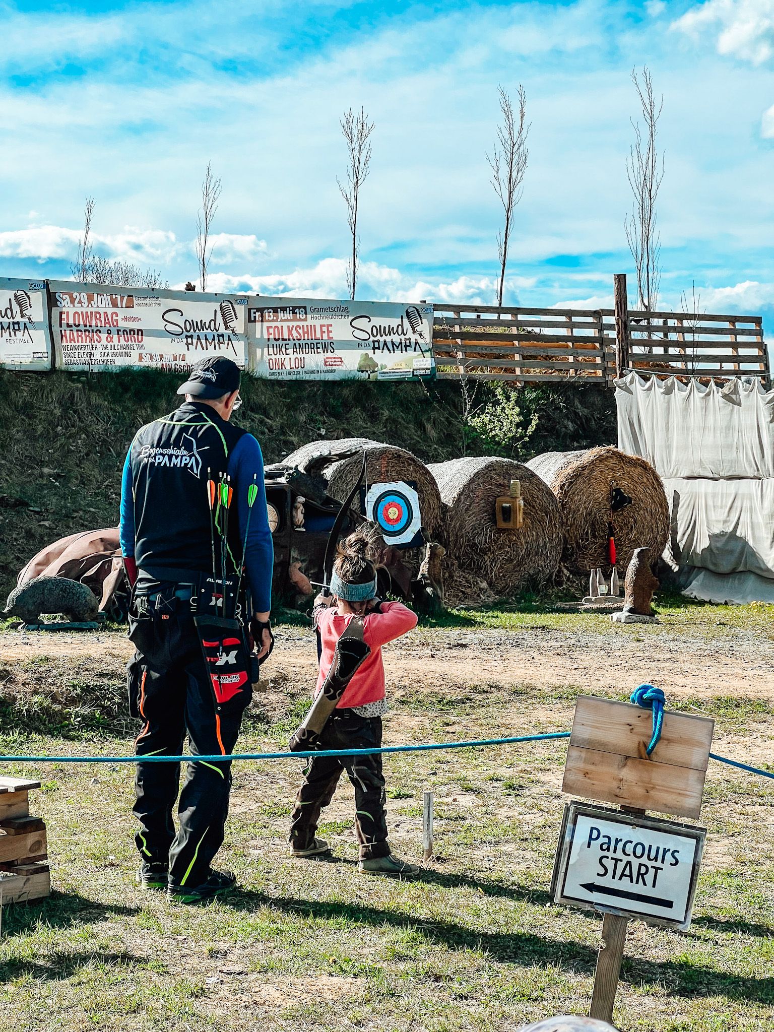 Dospelý a dieťa stoja na lukostreleckom ihrisku. V pozadí sú terče a nápis "Parcours START".