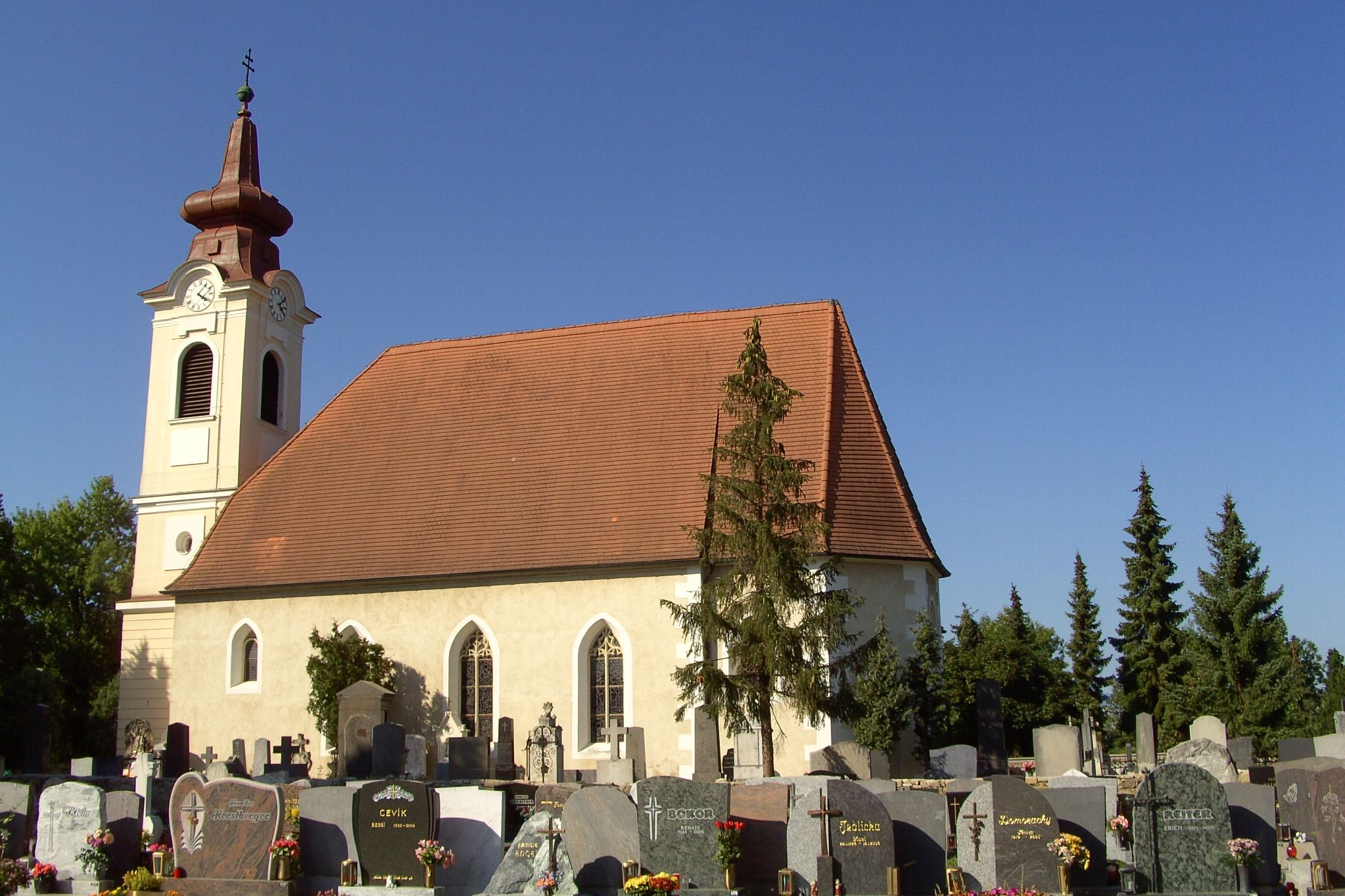 Exteriérový pohľad na kostol s cintorínom v popredí a modrou oblohou.