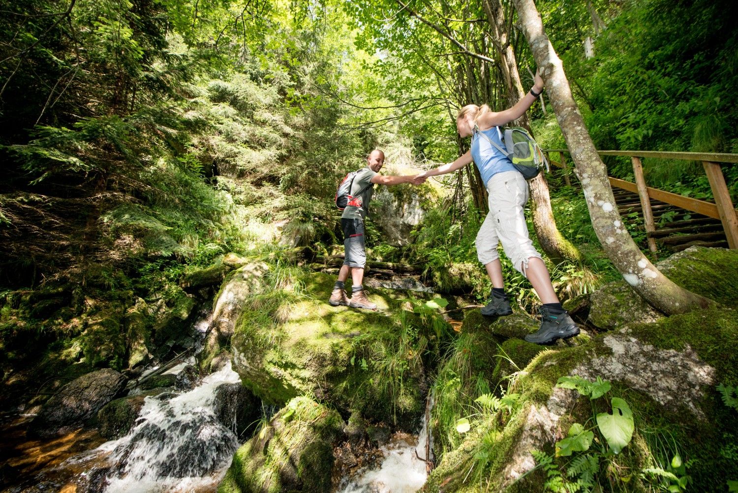 Dvaja turisti si navzájom pomáhajú prejsť cez potok v zelenom lese.