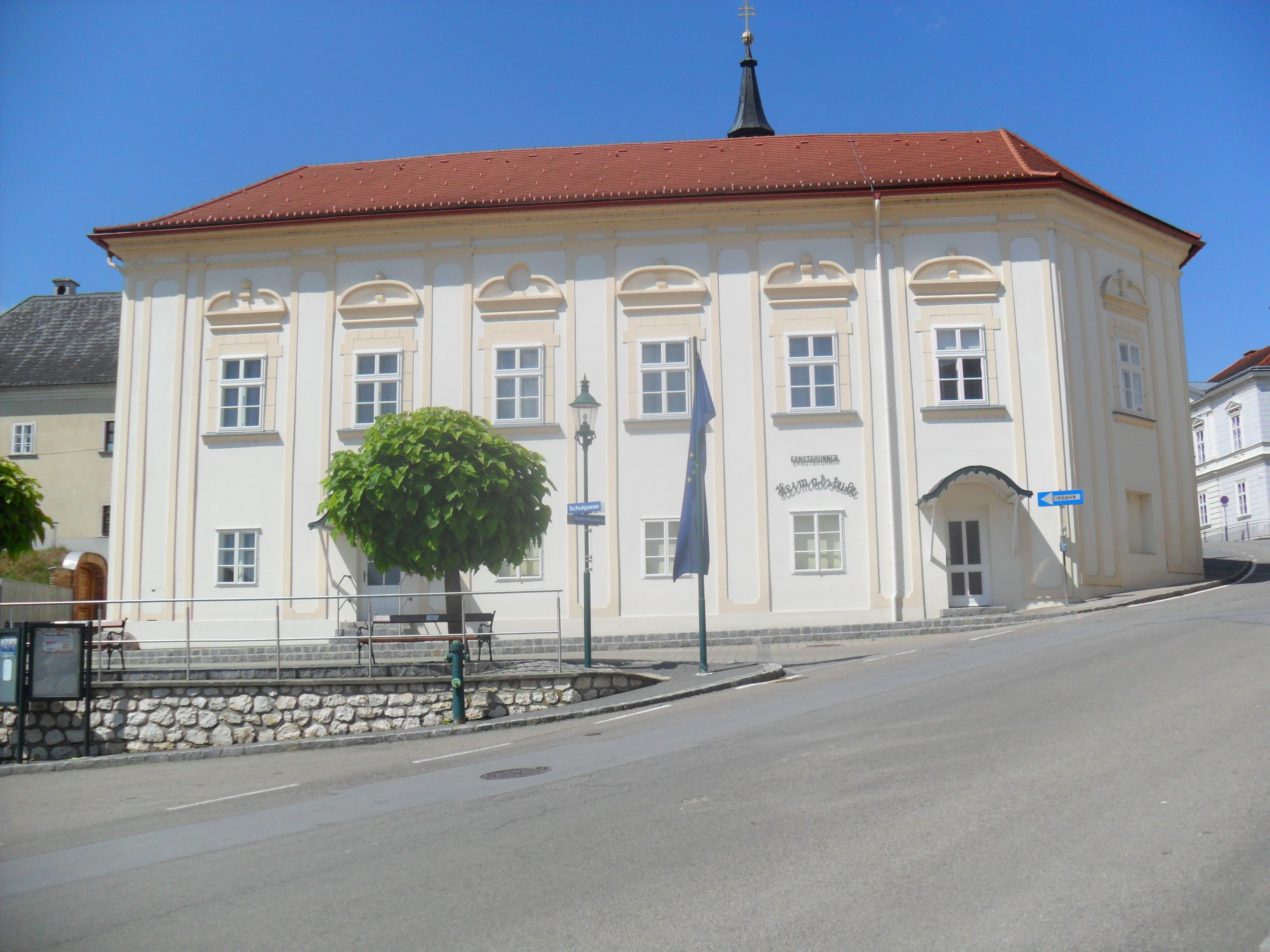 Historická budova s červenou strechou a bielymi stenami, obklopená stromami a cestou.