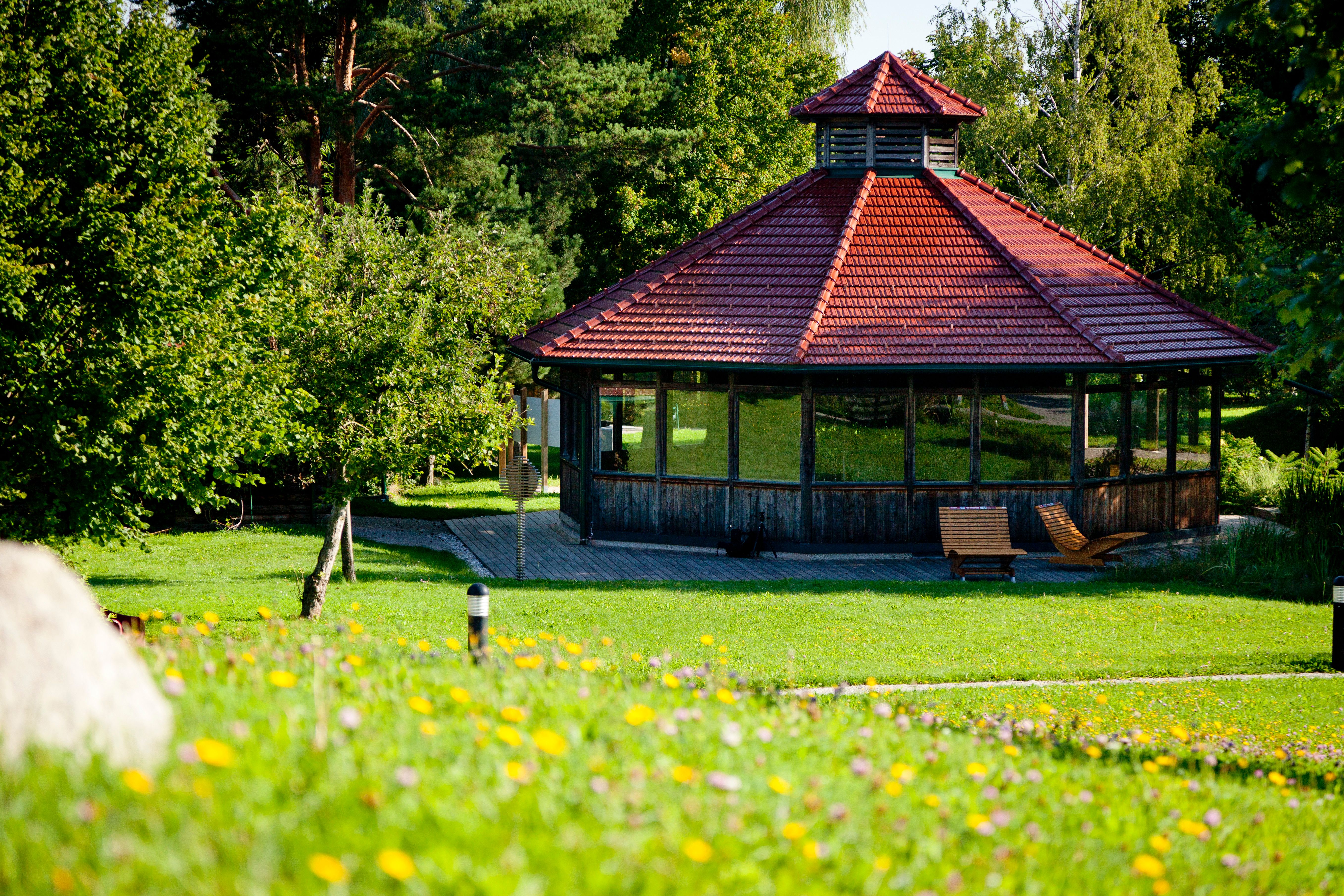 Pavilón s červenou strechou v zelenom parku, obklopený stromami a kvetmi.