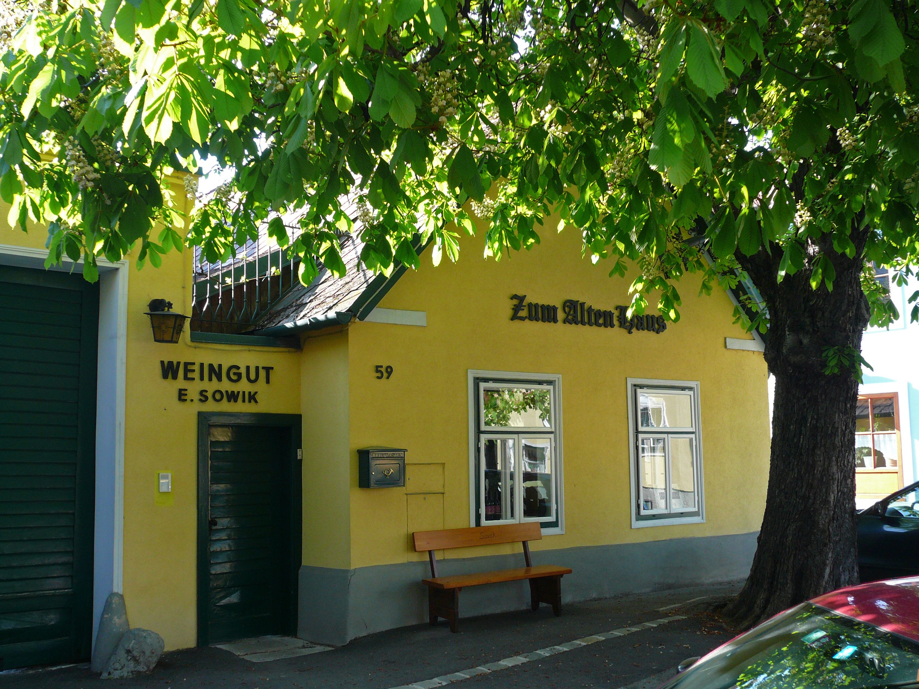 Žltá budova s nápismi "Zum Alten Haus" a "Weingut E. Sowik", obklopená zelenými stromami.