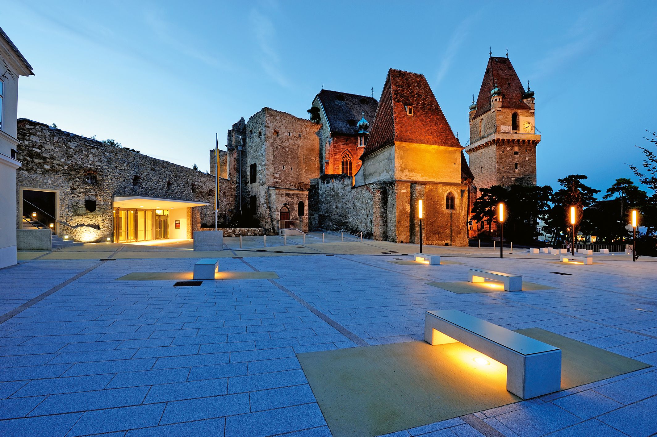 Večerný záber na osvetlený hrad Perchtoldsdorf s moderným predpolím a lavičkami.