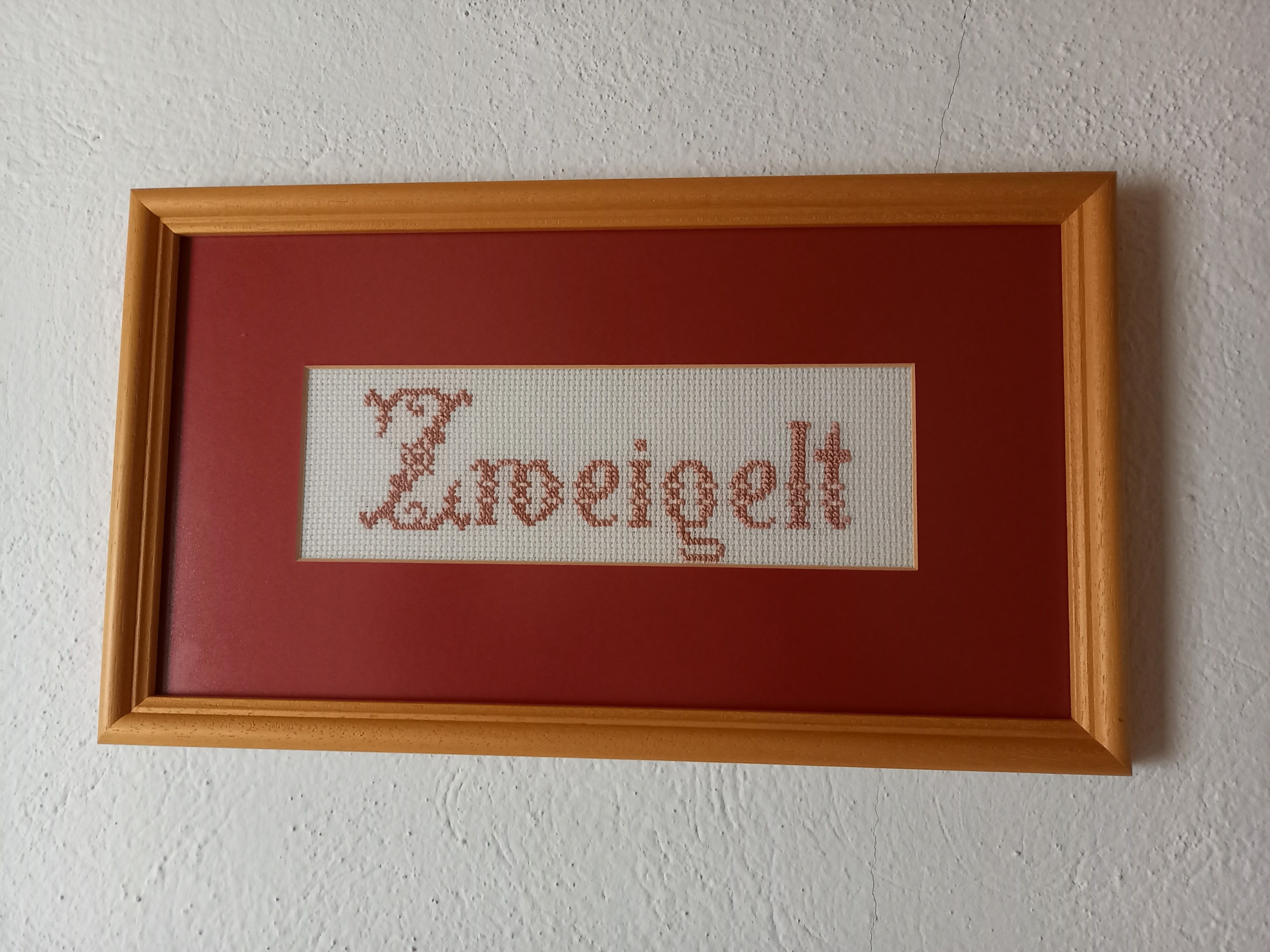 Vyšívaný obrázok s nápisom "Zweigelt" v drevenom ráme.
