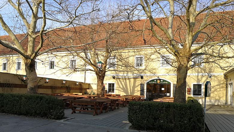 Žltá budova s červenými škridlami na streche a pivná záhrada s drevenými lavicami a stolmi v popredí.
