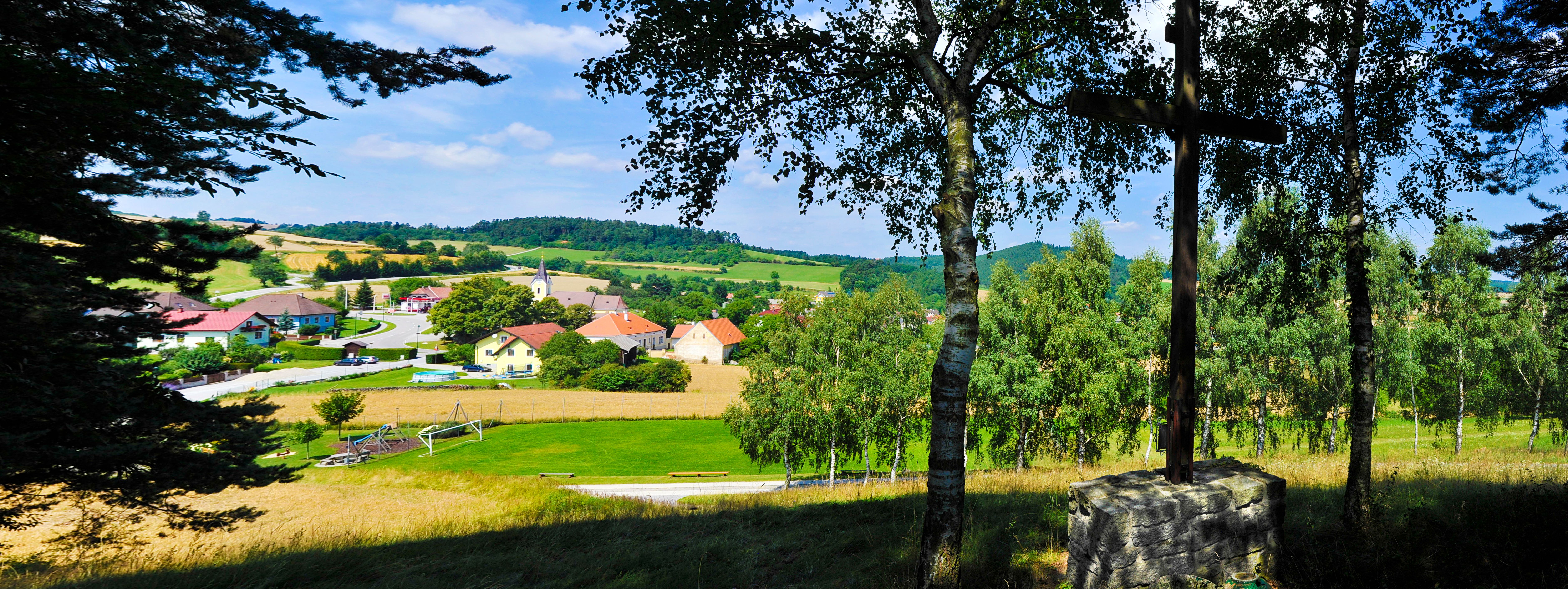 Pohľad na vidiecku obec s kostolom a domami, obklopenú poliami a stromami.
