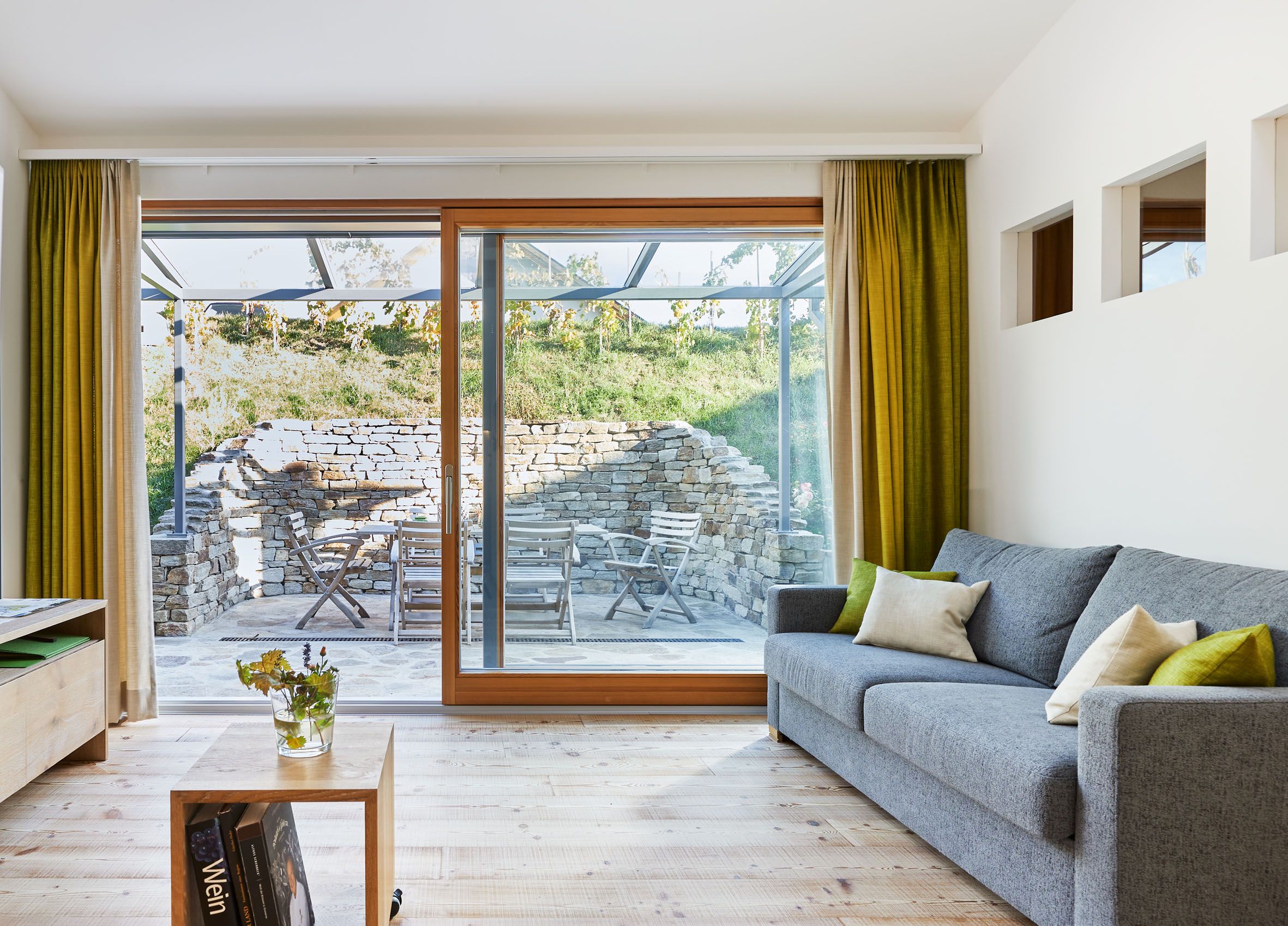 Moderná obývacia izba so sivou pohovkou, drevenou podlahou a veľkými oknami s výhľadom na terasu s kamennou stenou a záhradným nábytkom.