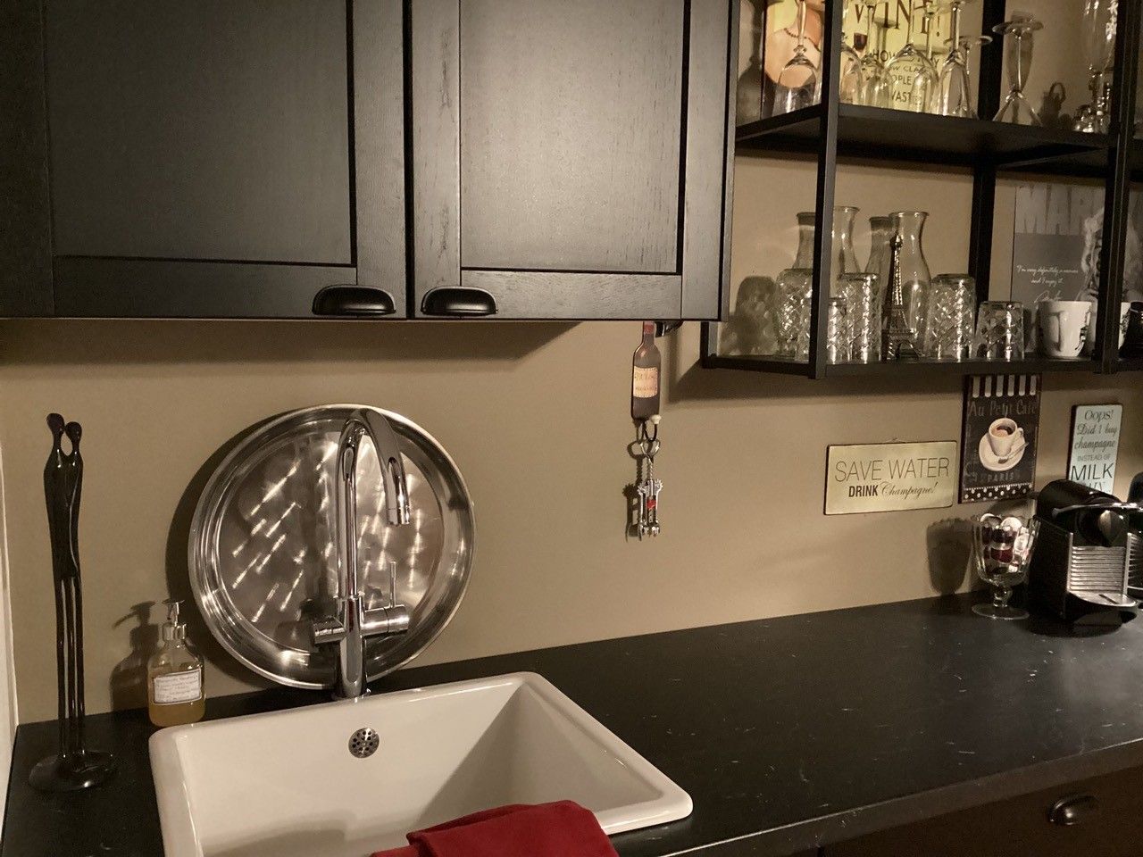 Moderný kuchynský kút s drezom, čiernou skriňou, policami s pohármi a kávovarom.