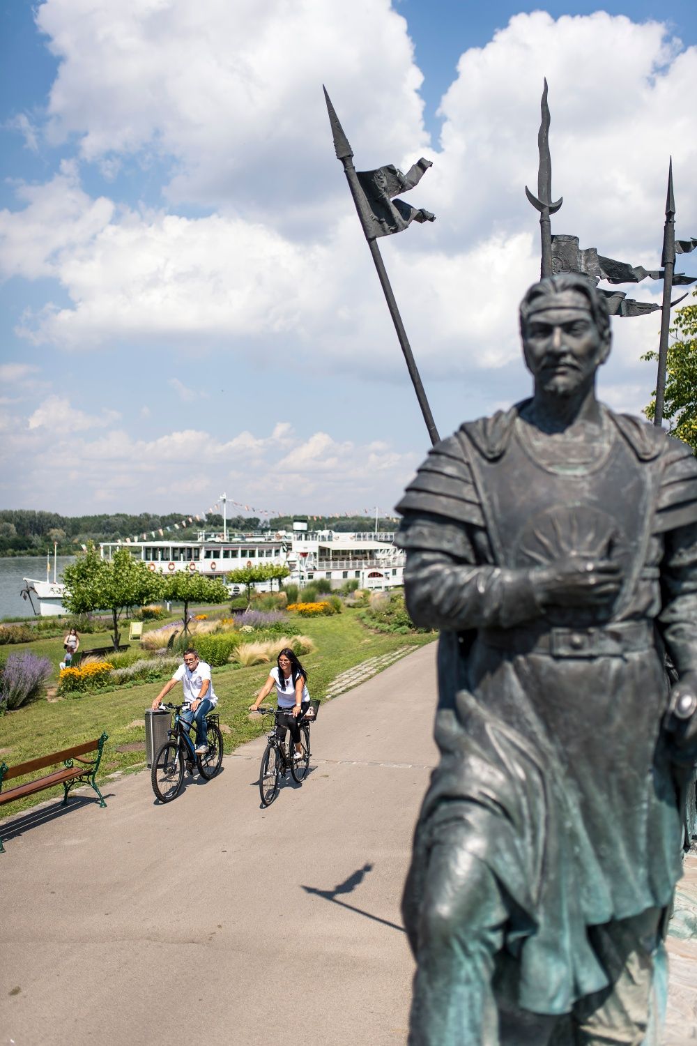 Dvaja cyklisti jazdia po ceste pozdĺž Dunaja v Tullne. V popredí je socha a v pozadí vidieť riečnu výletnú loď.