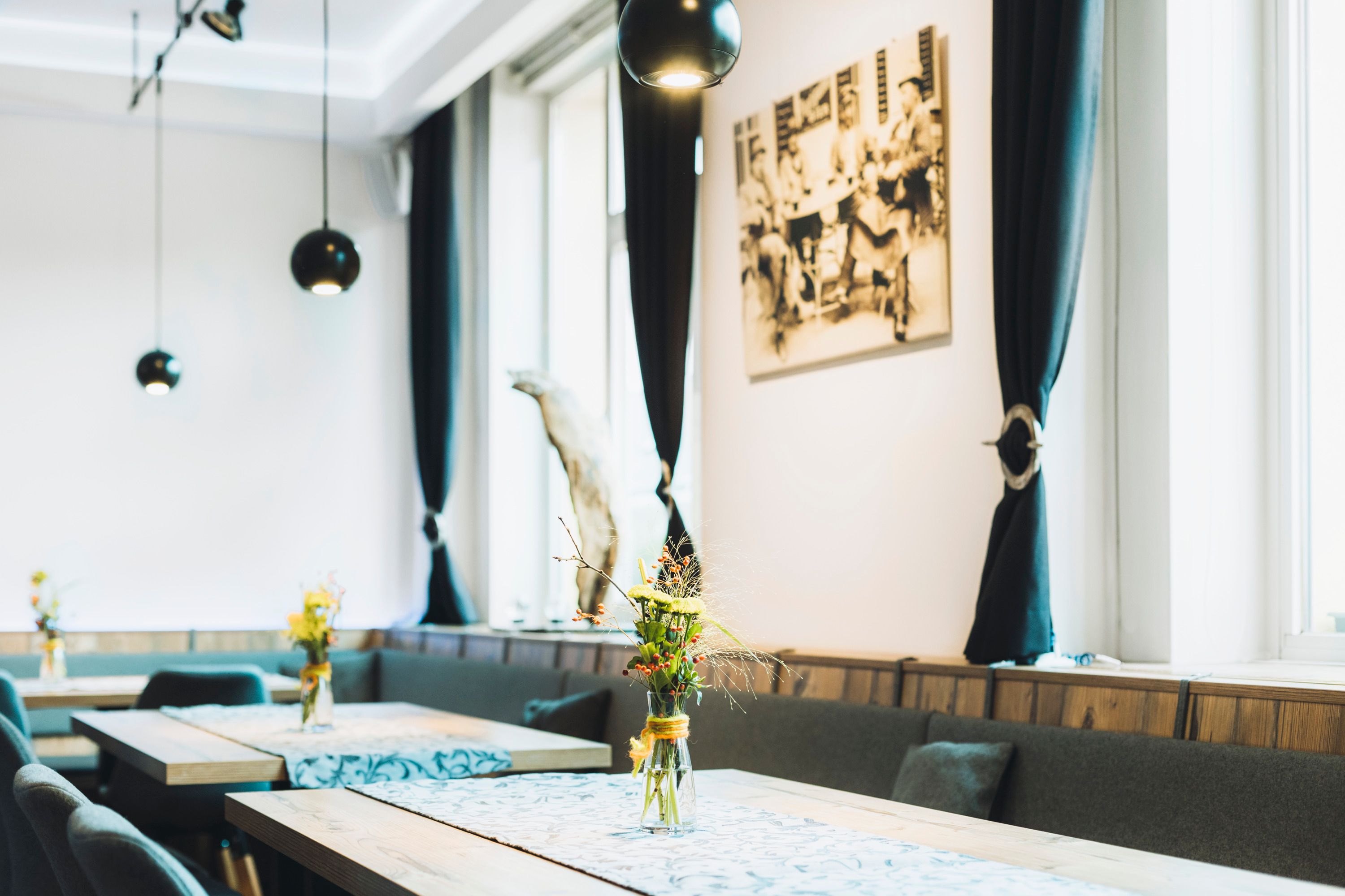 Pohľad do interiéru štýlovej kaviarne s drevenými stolmi, vázami na kvety a čiernymi závesmi.