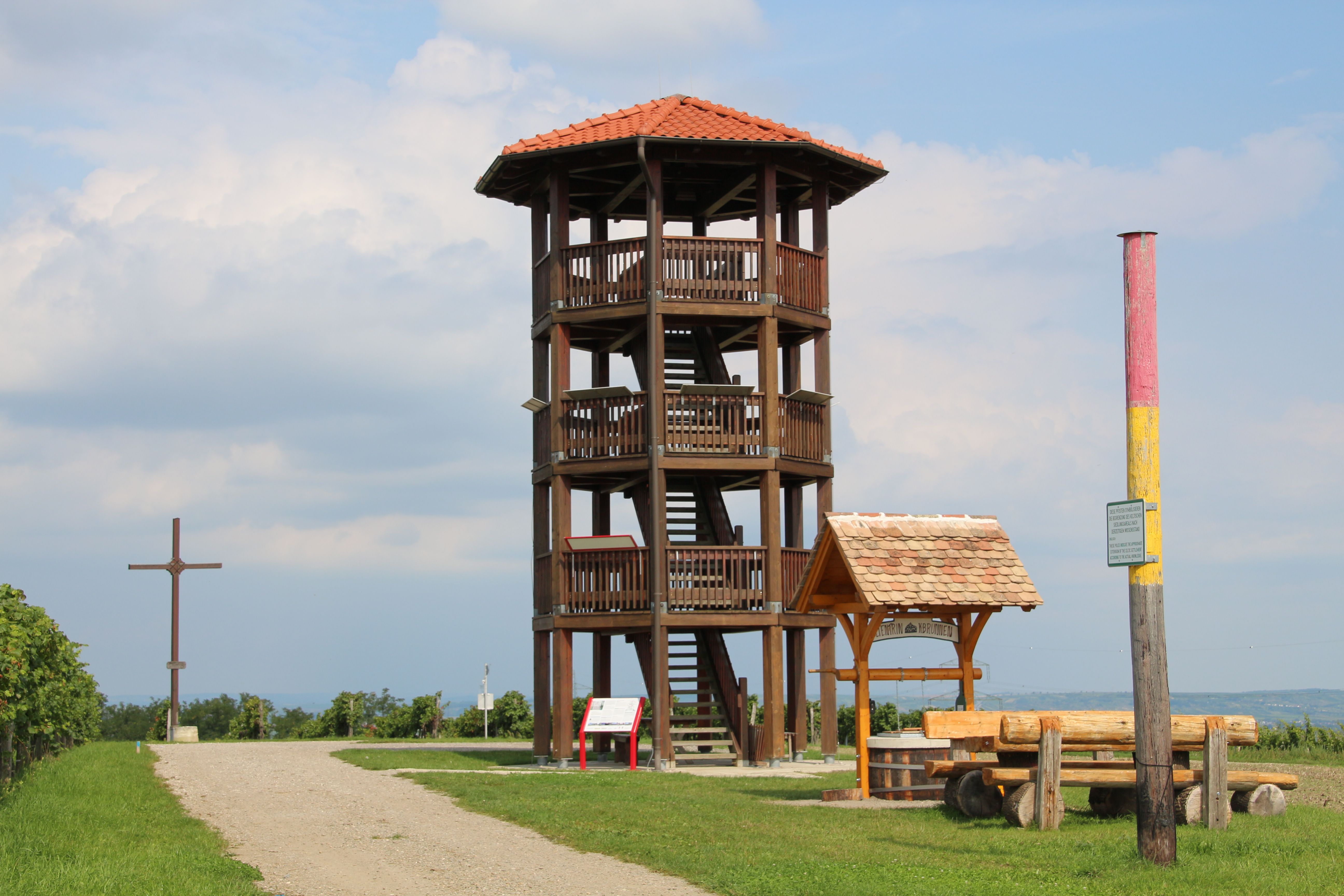Drevená rozhľadňa s červenou strechou na lúke, vedľa nej drevený kríž a lavička.