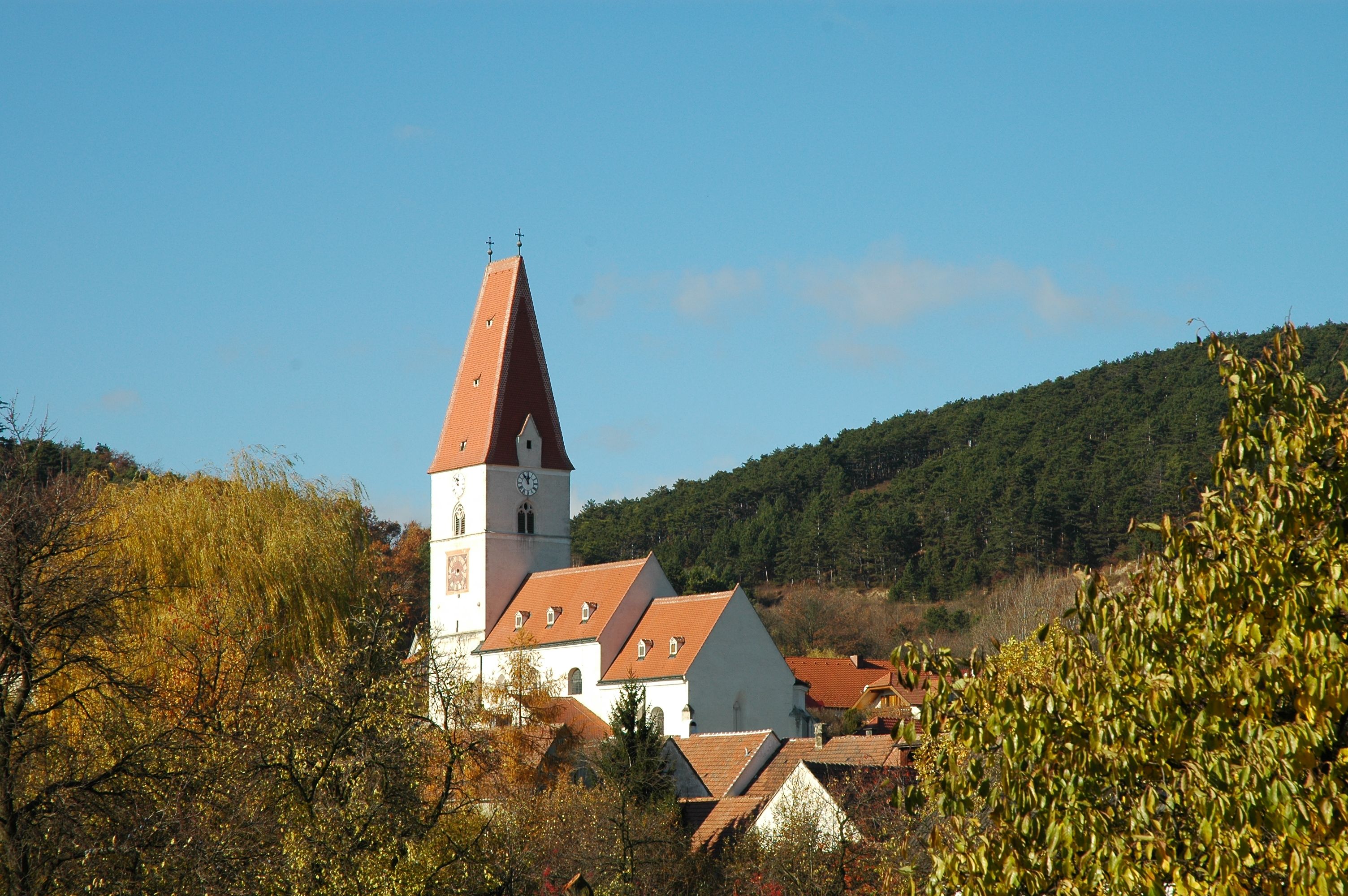 Kostol v Nußdorfe s červenou strechou a vežou s hodinami, obklopený stromami a kopcami.