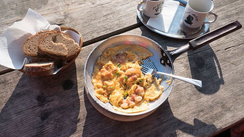 Panvica s miešanými vajíčkami a slaninou na drevenom stole, vedľa nej košík na chlieb a dva poháre.