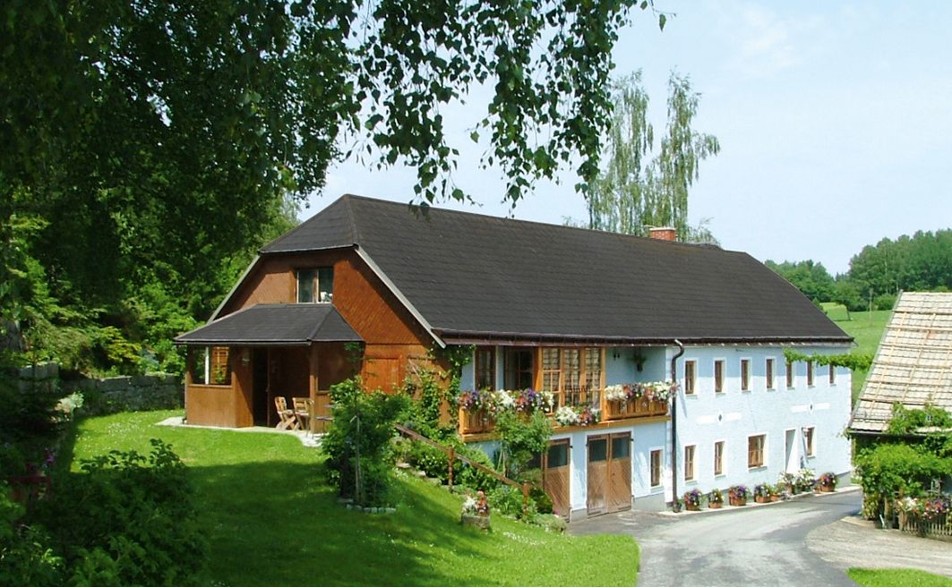Tradičný statok s kvetmi na balkóne, obklopený zelenou krajinou a stromami.