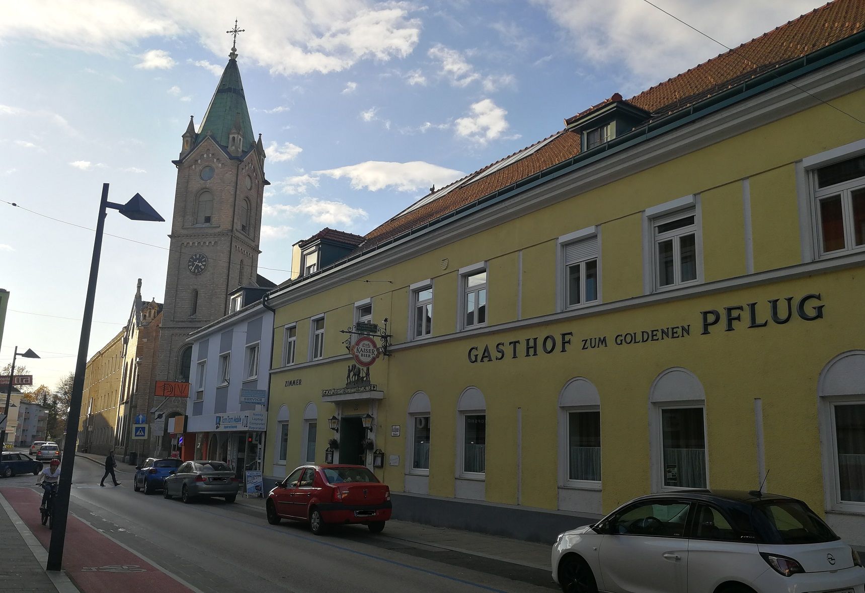 Scéna na ulici so žltou budovou "Gasthof zum goldenen Pflug" a kostolnou vežou v pozadí.