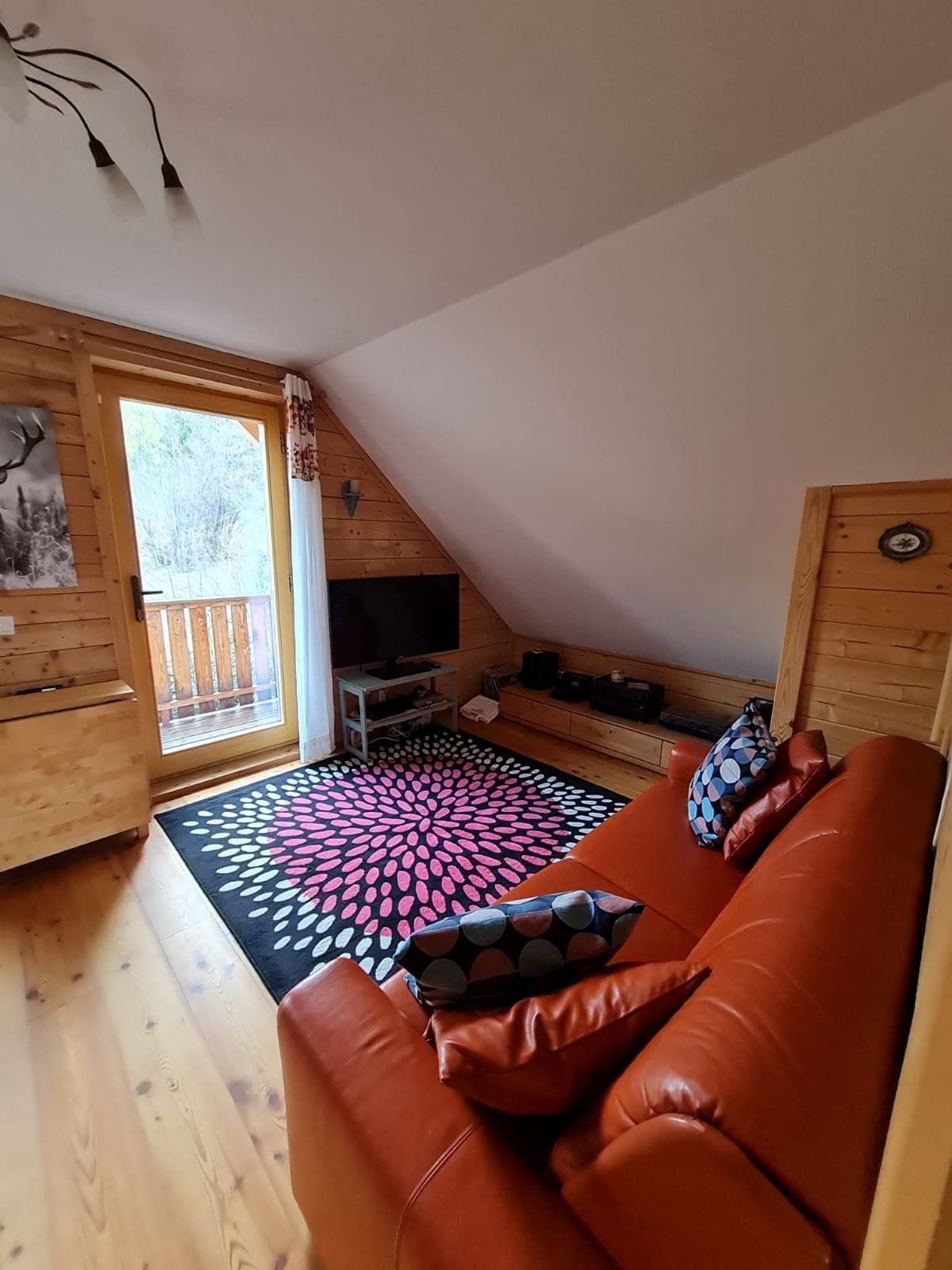 Útulná obývacia izba s červenou pohovkou, drevenými stenami a farebným kobercom pred balkónovým oknom.