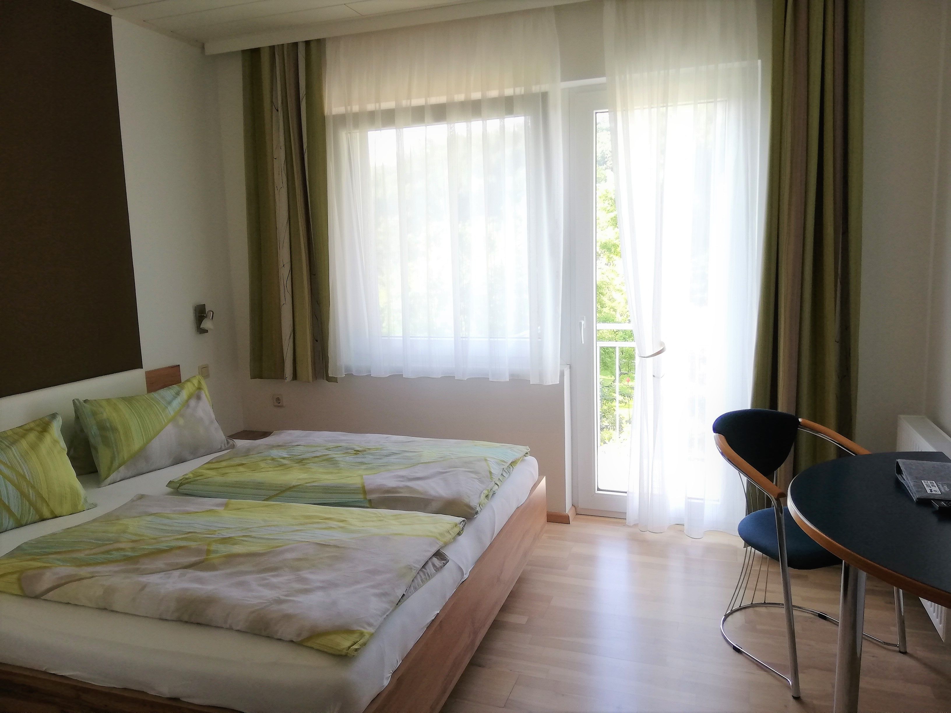 Dvojlôžková izba s posteľou, stolom a stoličkou. Cez sklenené dvere je viditeľný balkón.