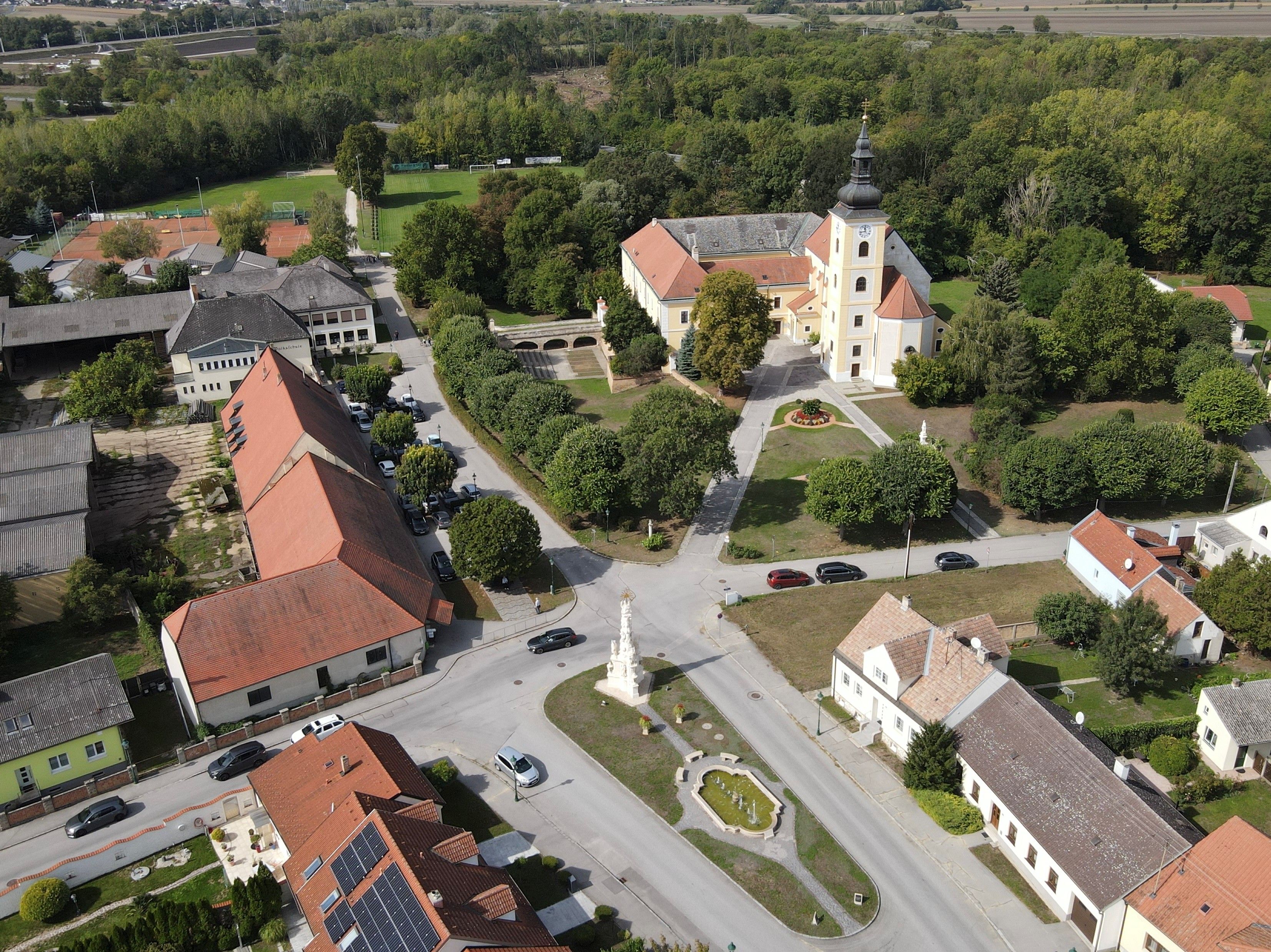 Letecký pohľad na Weikendorf s kostolom, námestím, fontánou a okolitými domami na okraji lesného porastu.
