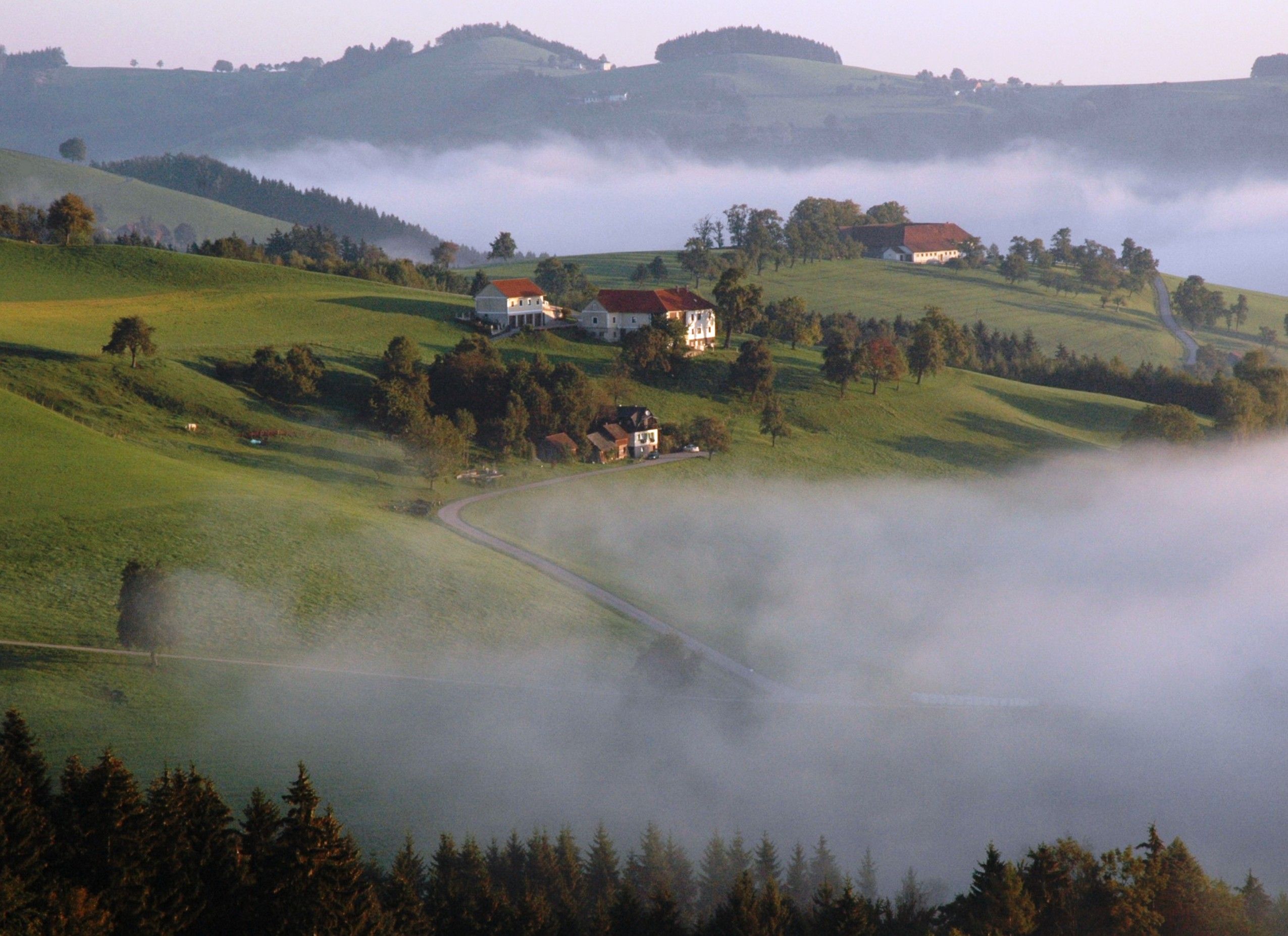 Jesenná krajina so zelenými kopcami, roztrúsenými domami a hmlou.