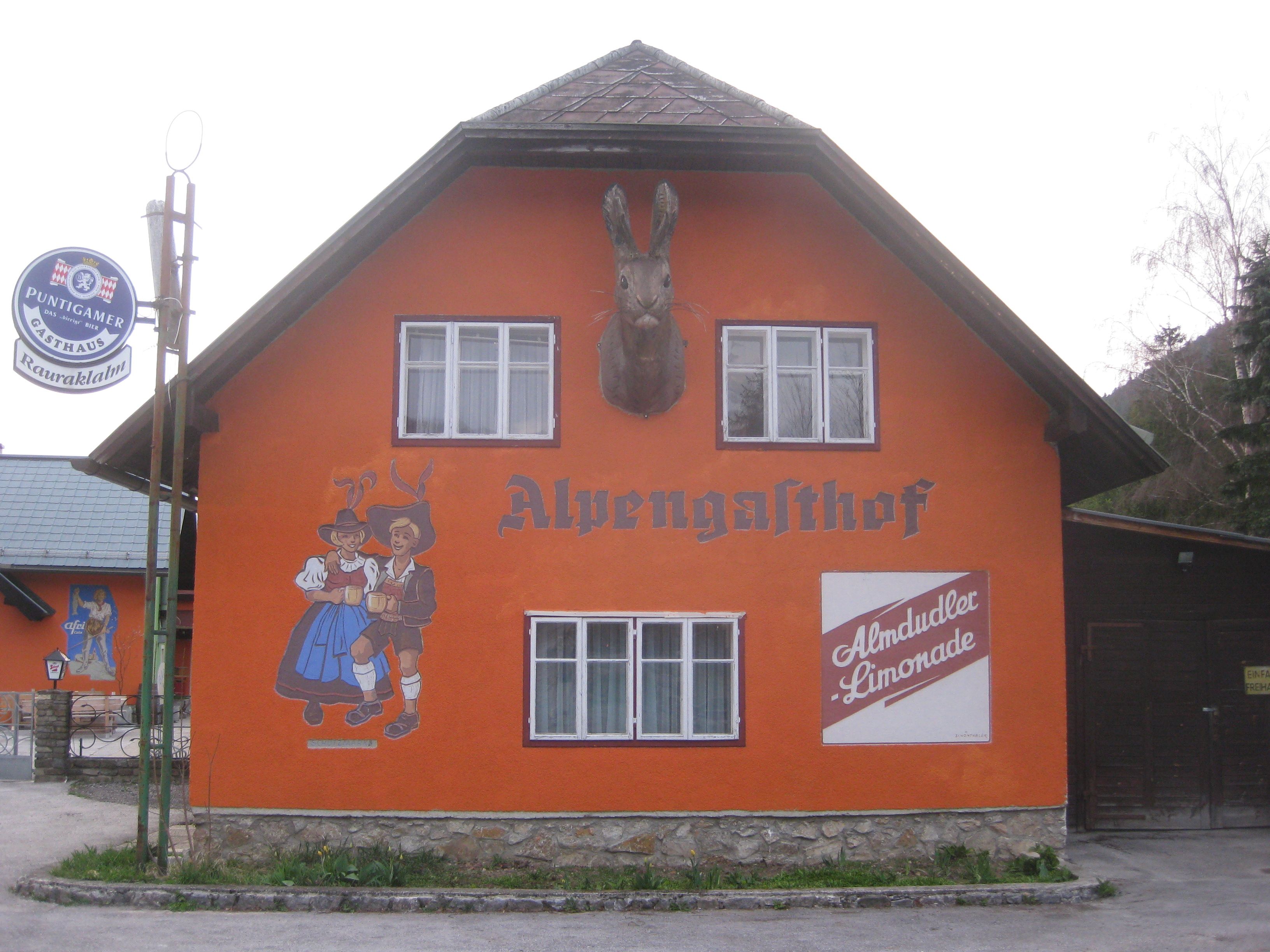 Oranžovo natretá budova s nápisom "Alpengasthof", nástenná maľba tancujúceho páru a reklama Almdudler.