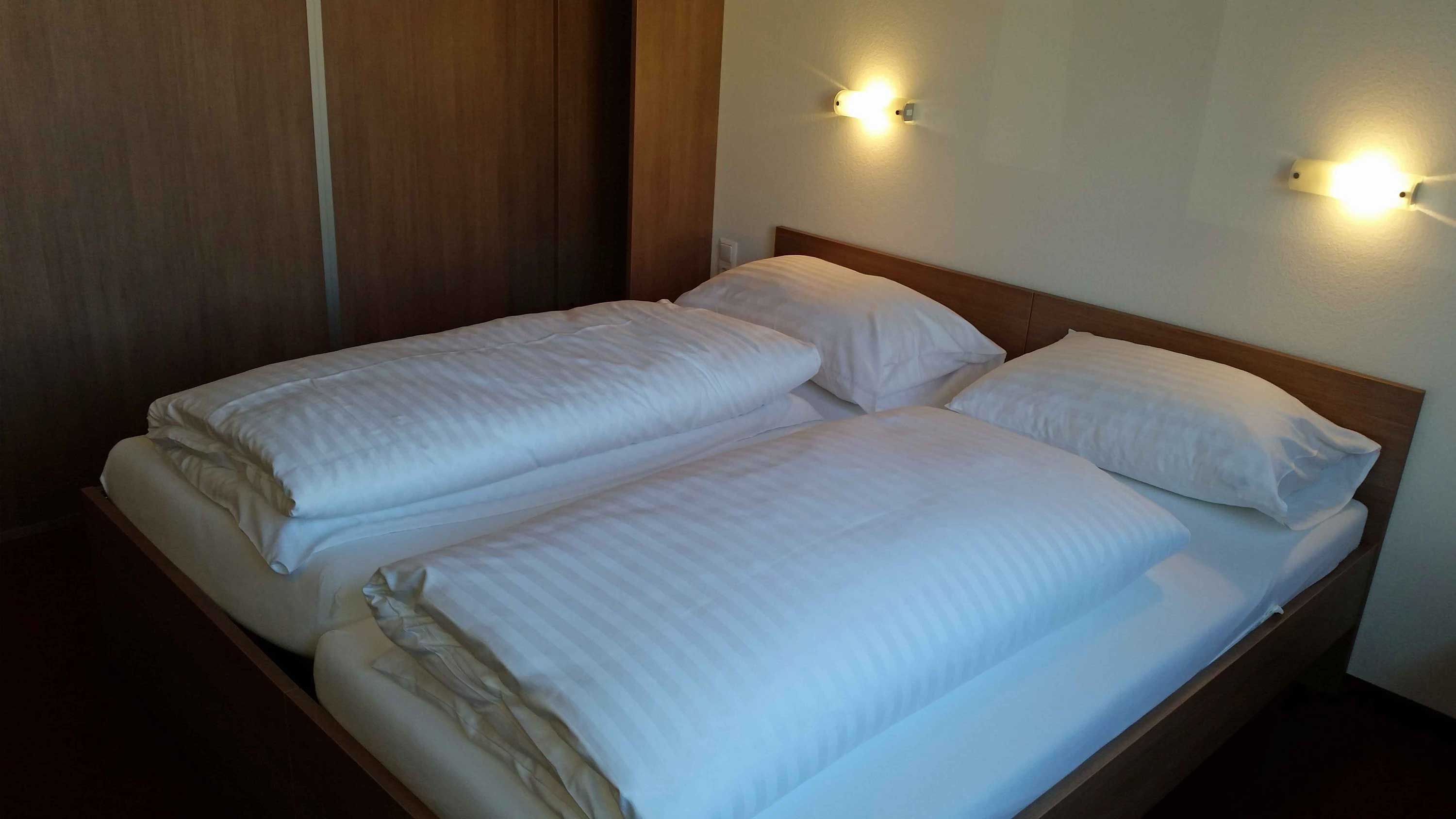 Manželská posteľ v motelovej izbe s bielymi perinami a vankúšmi, osvetlená dvoma nástennými lampami.