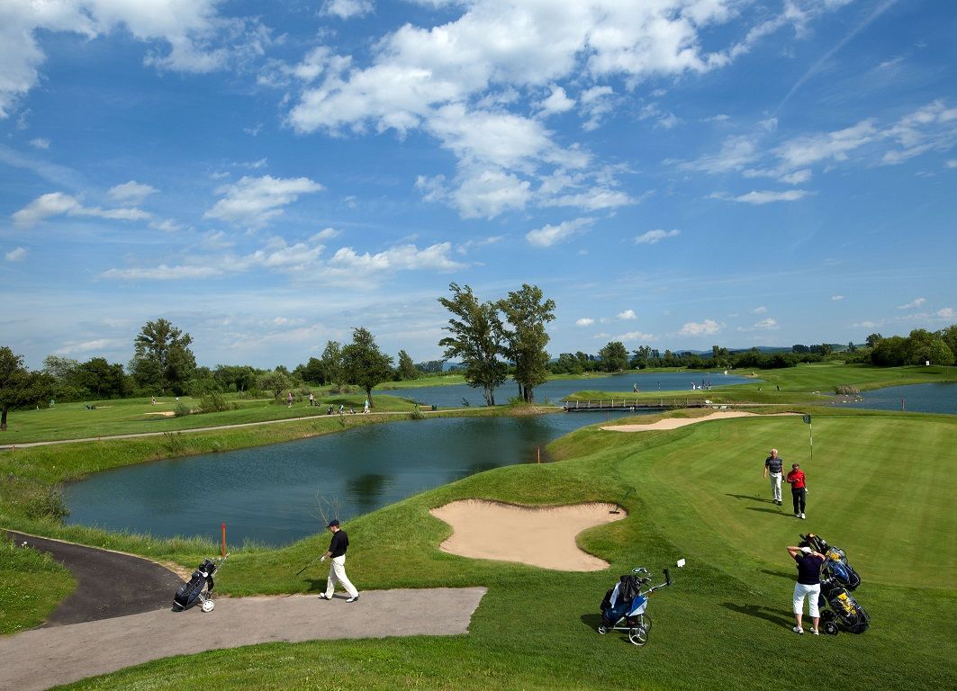 Golfové ihrisko s vodnými prekážkami a hráčmi pod modrou oblohou.