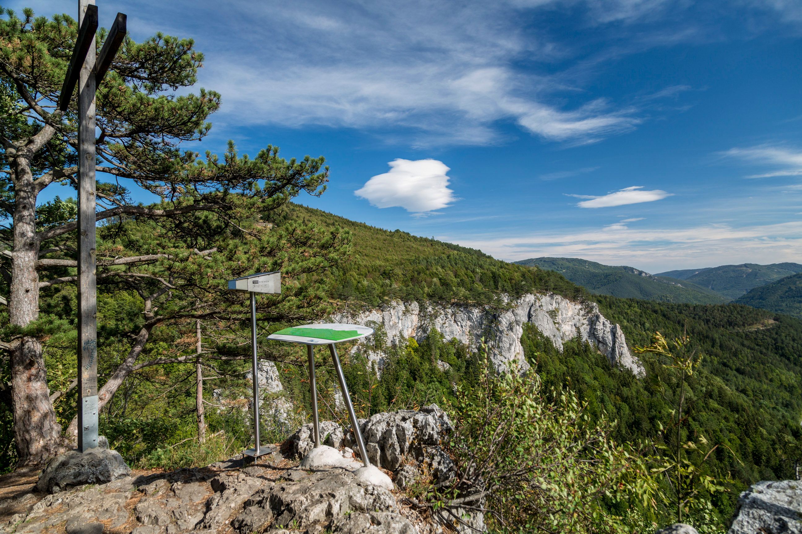 Vyhliadka s informačnou tabuľou a krížom na skale, obklopená lesom a horami pod modrou oblohou.
